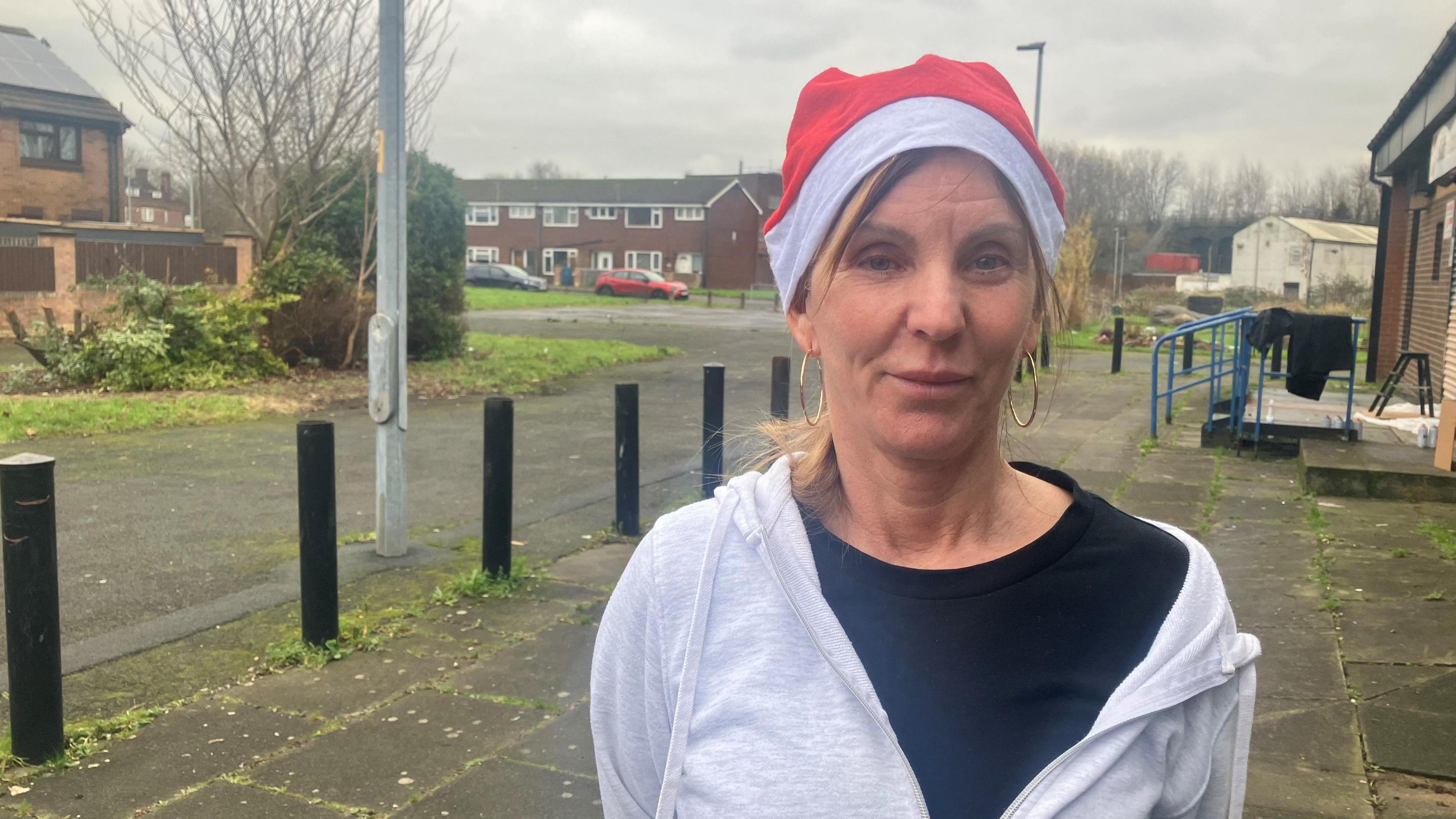 Photograph of Sharon Leech standing in the street in Collyhurst. She wears a dark blue t-shirt with a white hoodie. Smiling for the camera, she also wears a red-and-white Santa hat.