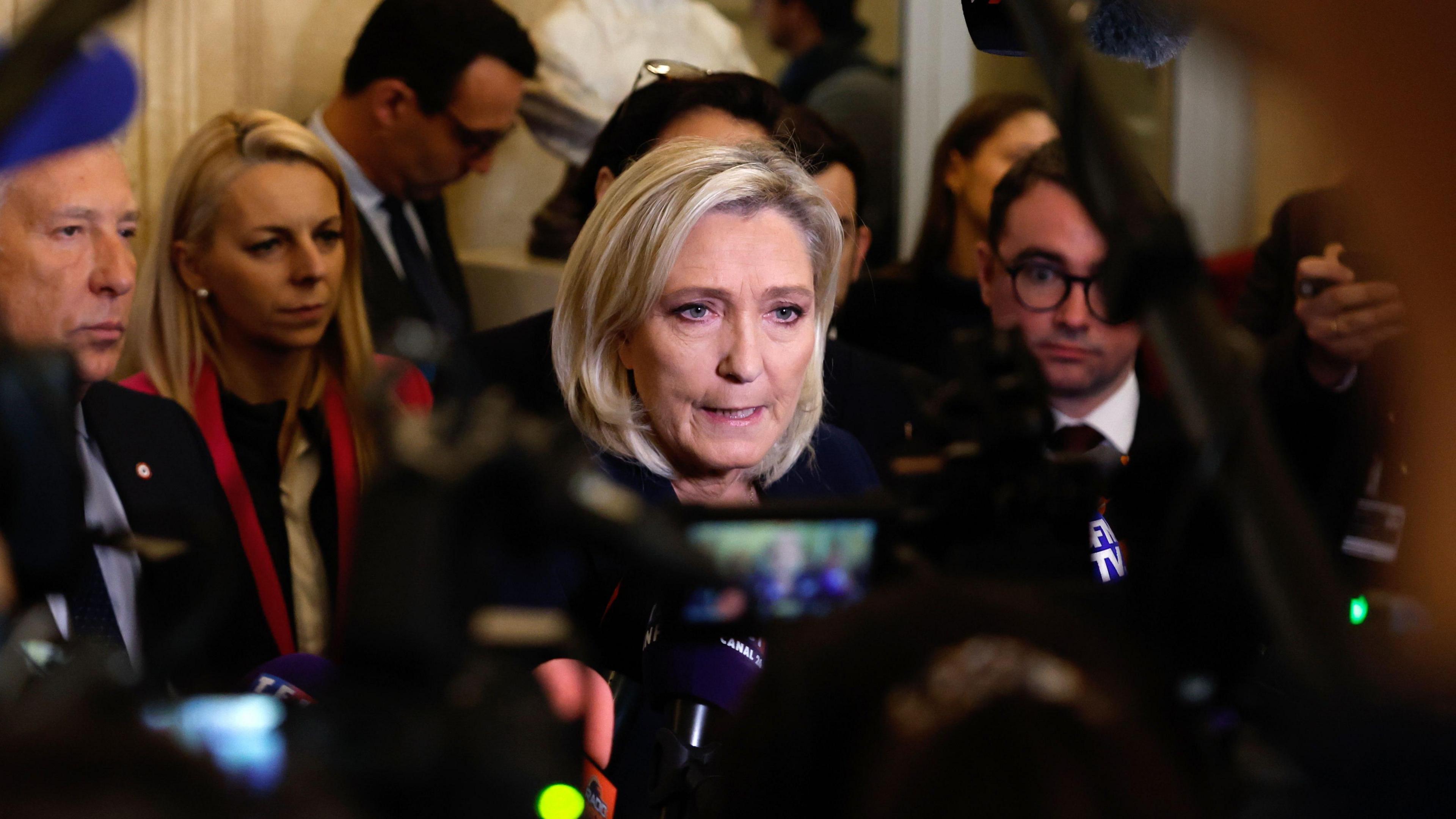 Marine le Pen, who has blonde bob and kohl-rimmed eyes, is surrounded by cameras as she speaks to journalists after the French National Assembly debate