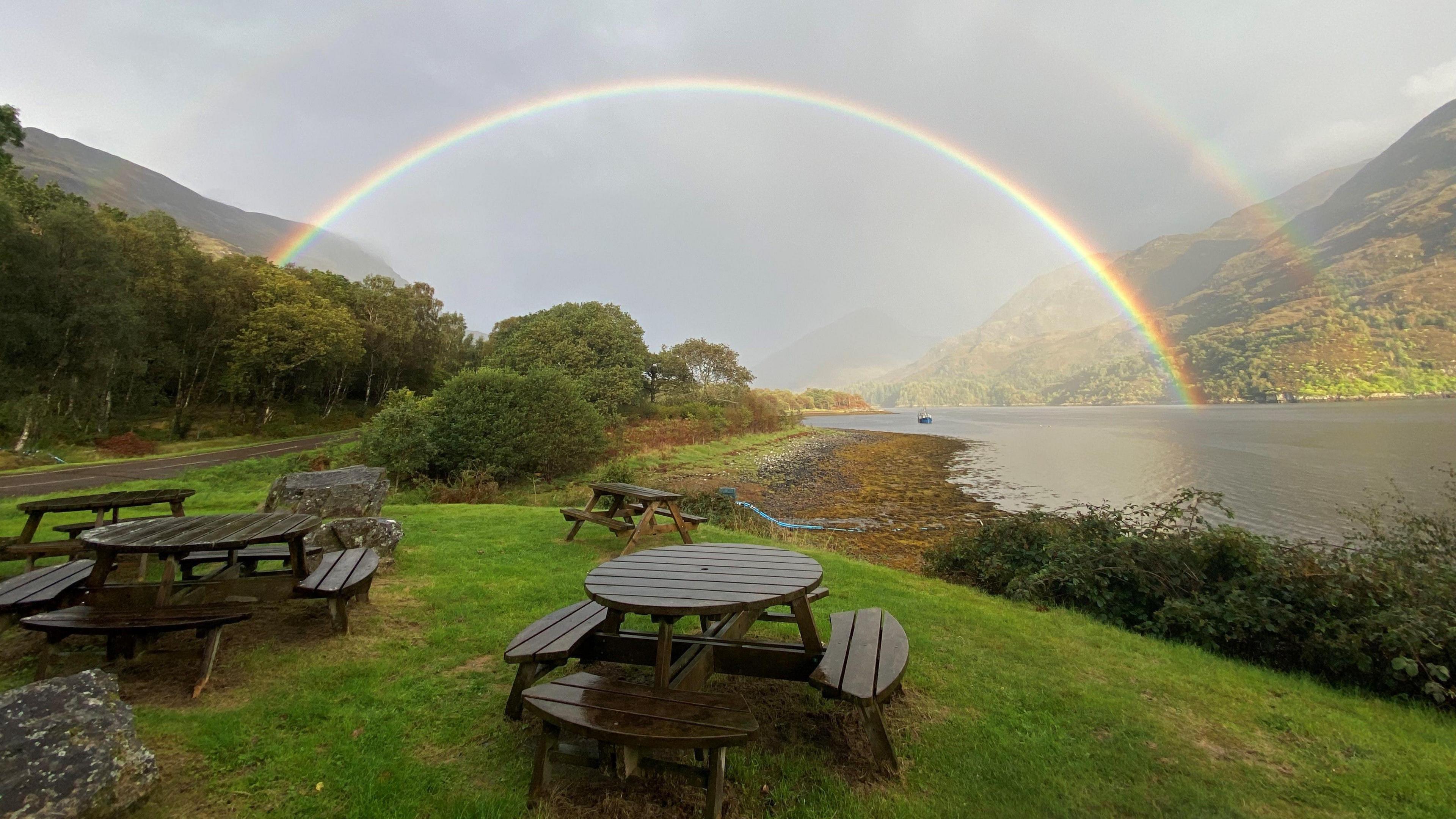 A double rainbow over a loch against a grey sky. There are circular picnic tables in the foreground on a green grass verge.