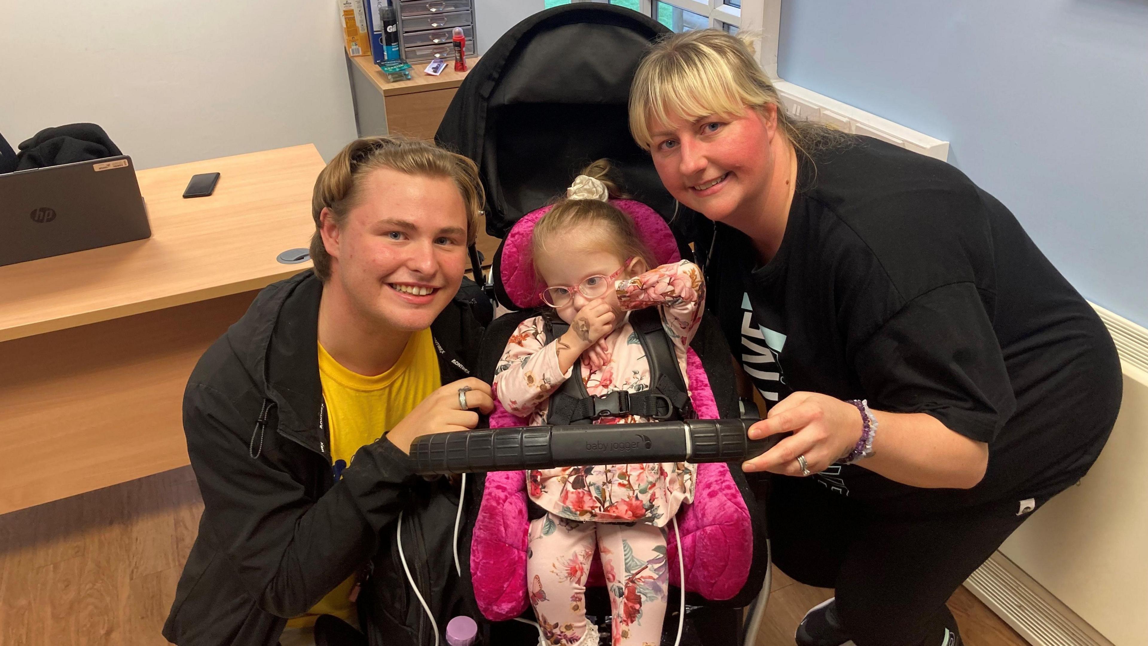 Three-year-old Robyn, with pink glasses and a pink flowery outfit, sits in a pram next to her smiling brother Josh, who is on the left and has blonde hair pushed back with a hairband, and her mother Stephanie Perry, who has a black t-shirt and blonde hair in a fringe