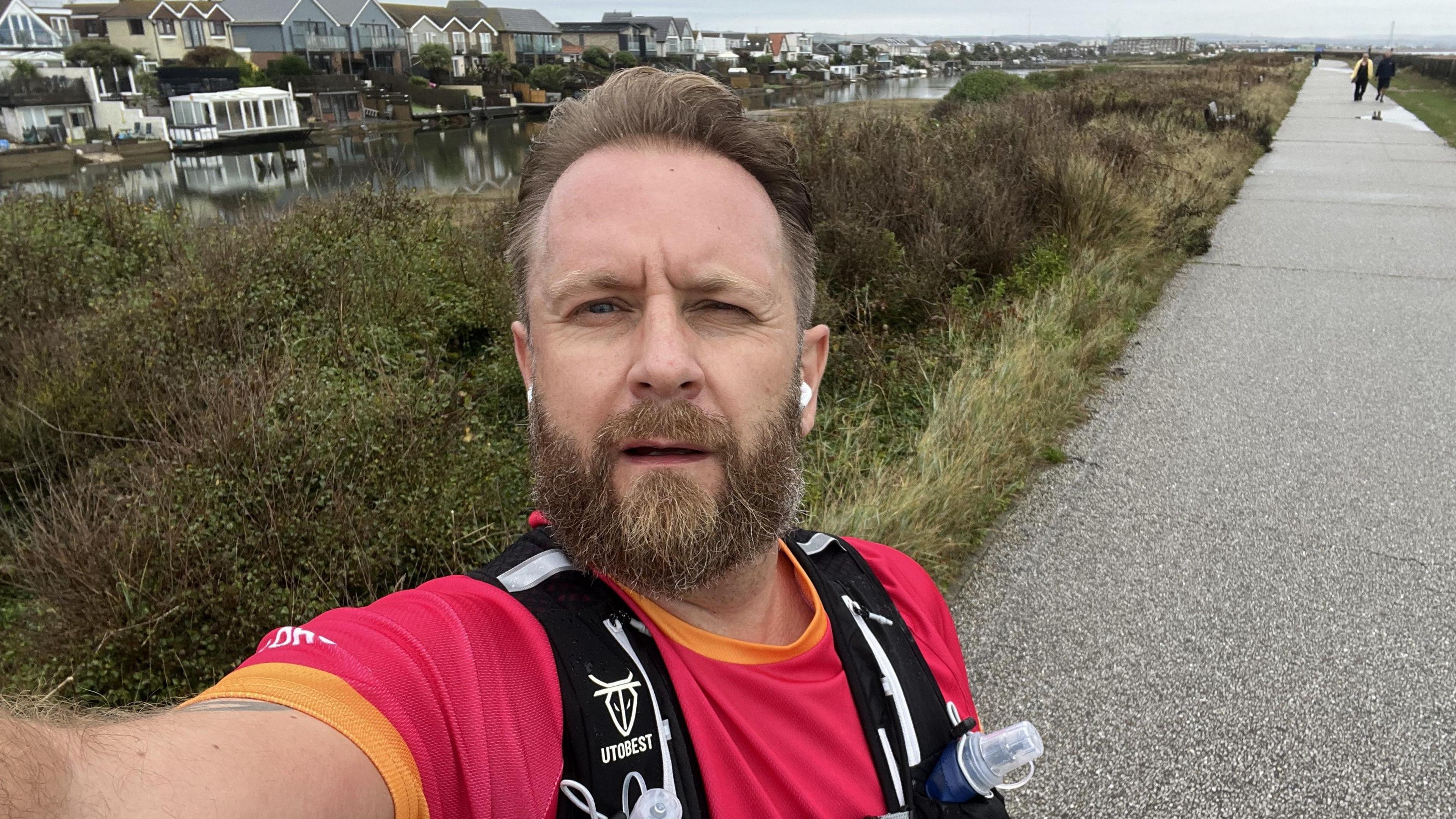 A bearded blond man takes a selfie on a canal path. He is wearing a red t-shirt and a running vest. 