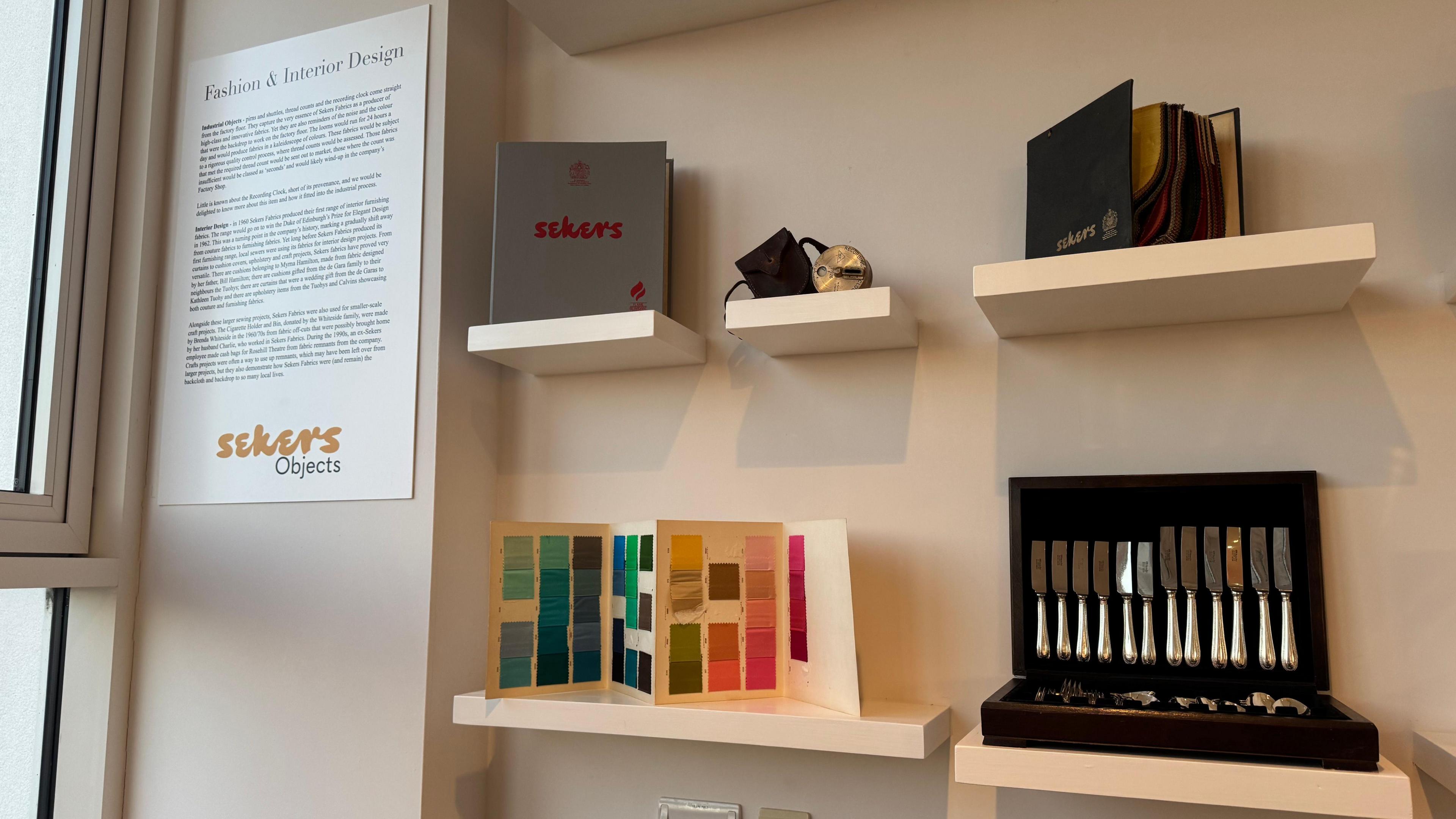 Some of the objects on display in an exhibition about Sekers are shown on different shelves. They include a box of knives and forks, some colour samples and a book of carpet squares.