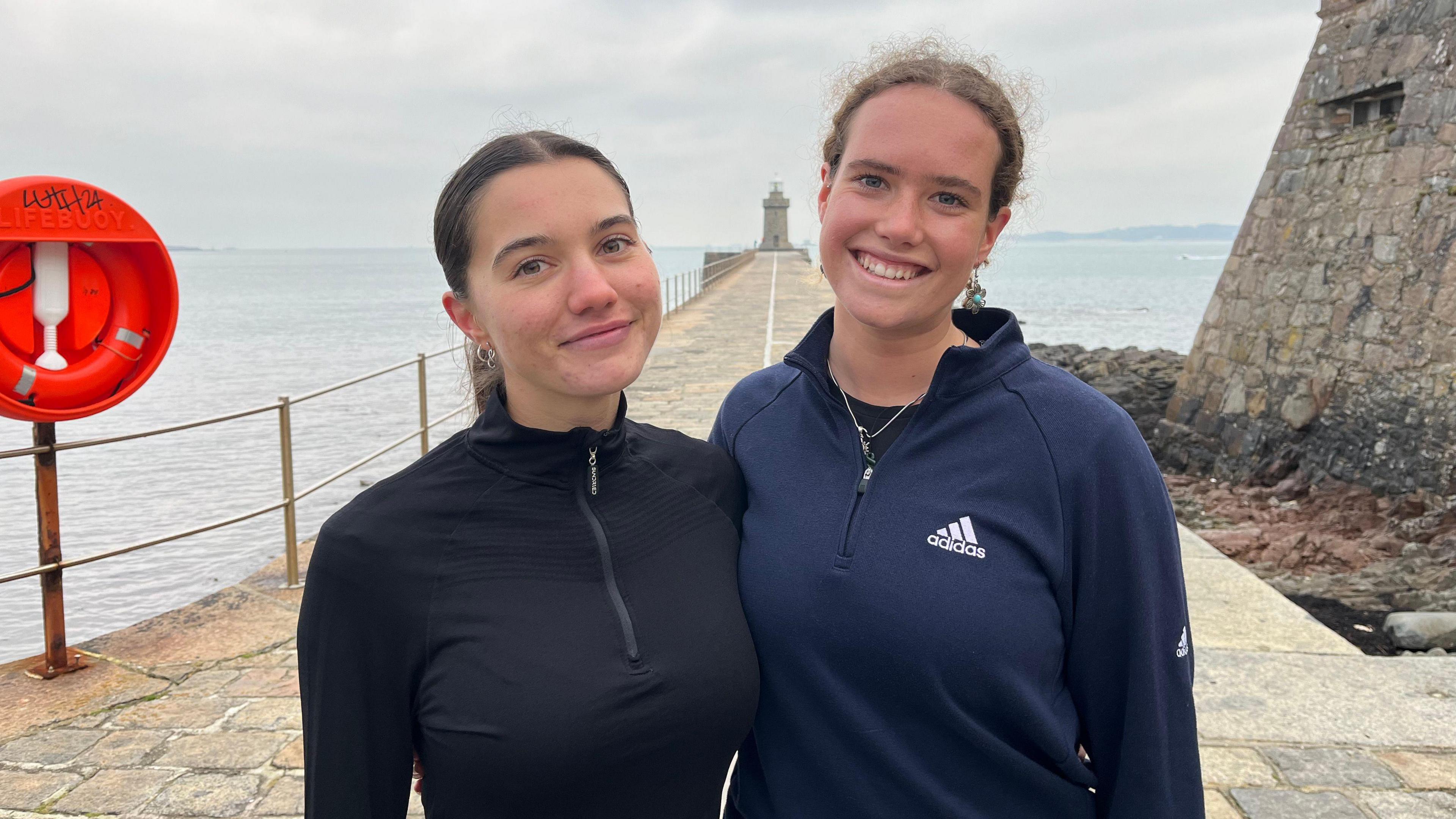 Lois Vidamour and Jenny Thomas smiling at the St Peter Port lighthouse. They are dressed in running quarter zips and their hair is tied back. 