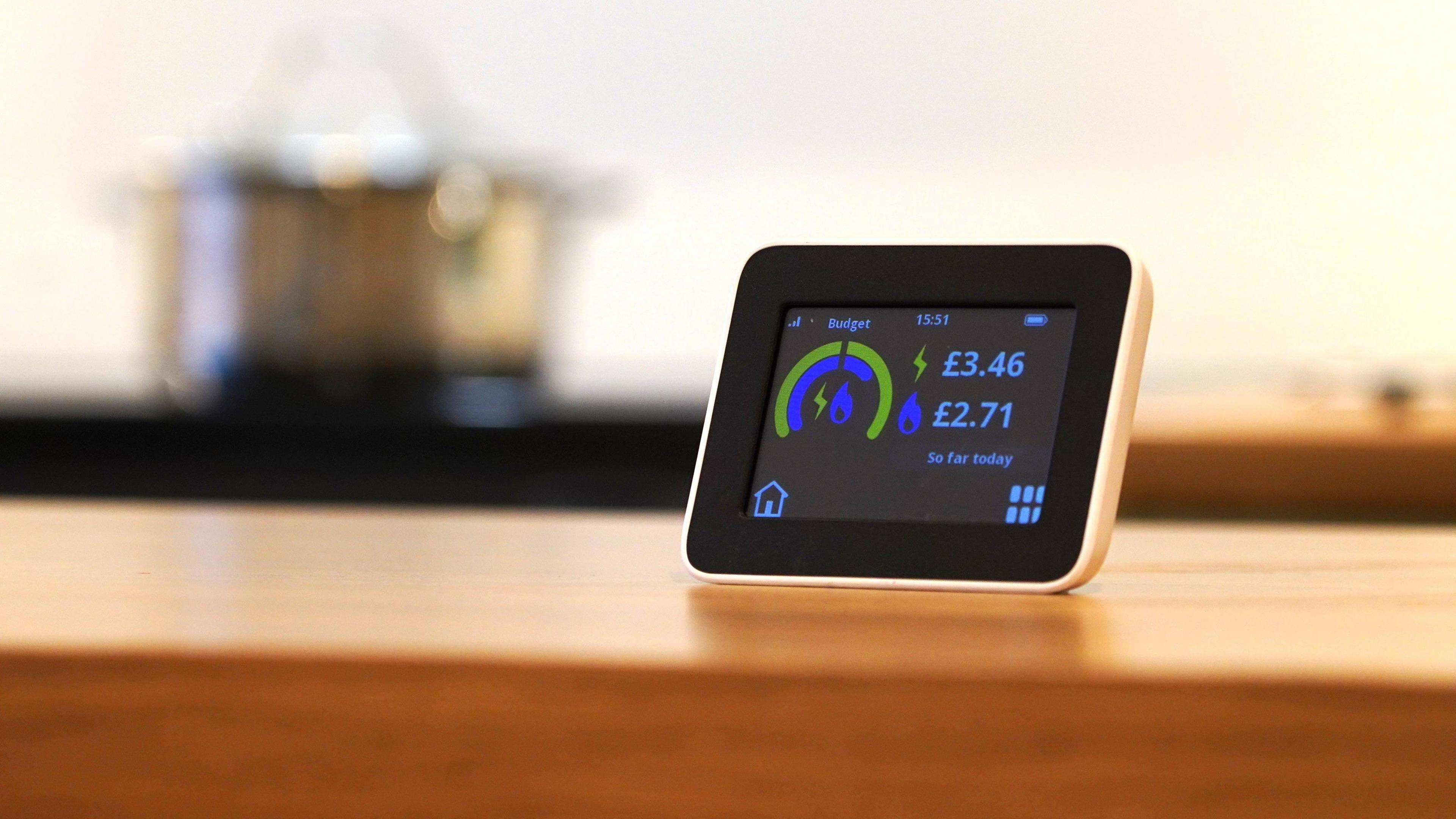 A picture of a small black smart meter on a wooden surface. The box has a yellow and blue semi-circle which highlights the electric and gas usage. Next to a bolt it says '£3.46' and a flame symbol with '£2.71' next to it. Underneath it says 'so far today'. In the bottom left corner there is a house symbol. 