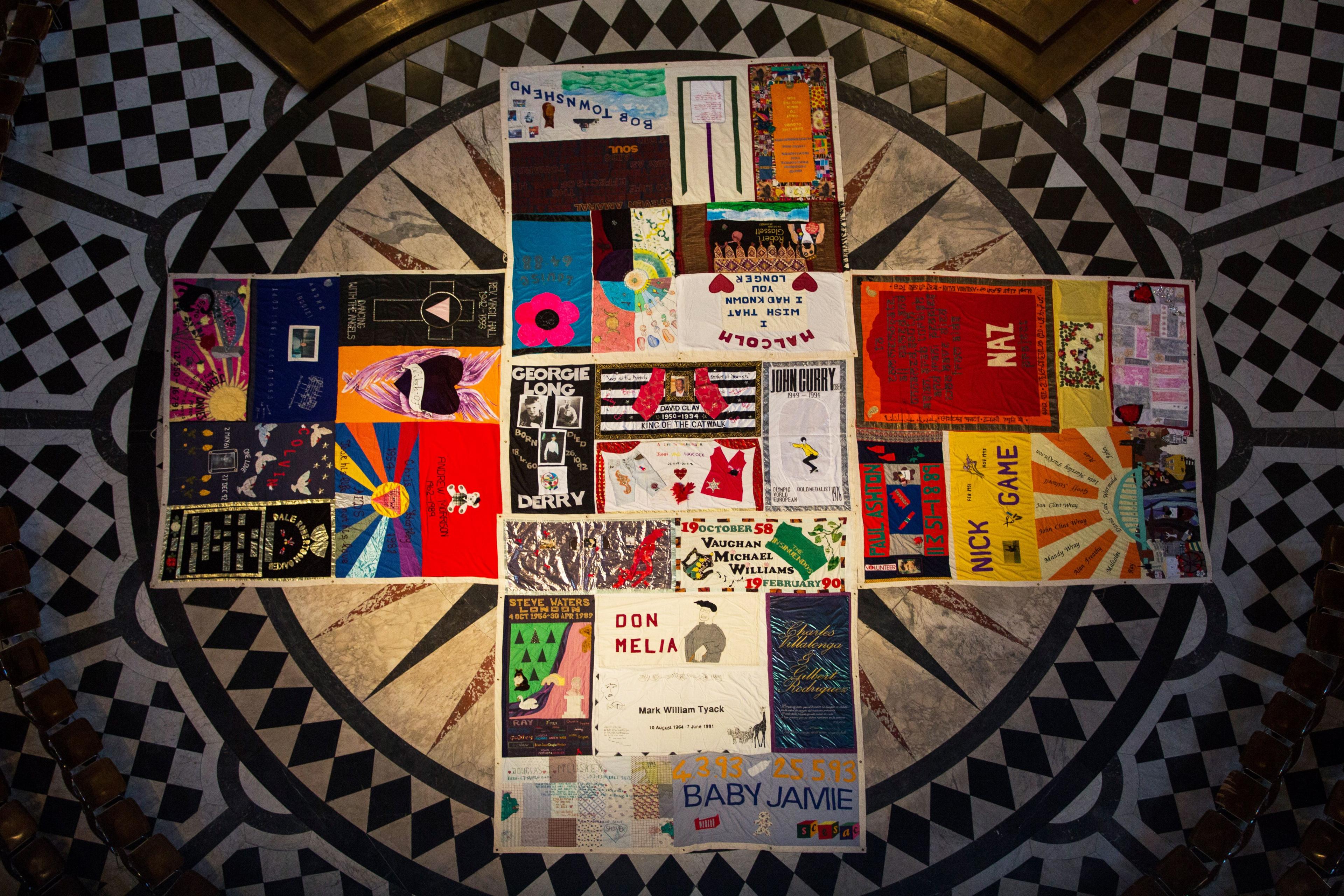 A portion of the UK AIDS Memorial Quilt on display at St Paul's Cathedral. The panels are arranged in the shape of a cross on the church floor. It is full of bright colours, and on it can be seen names like Nick and Malcolm.