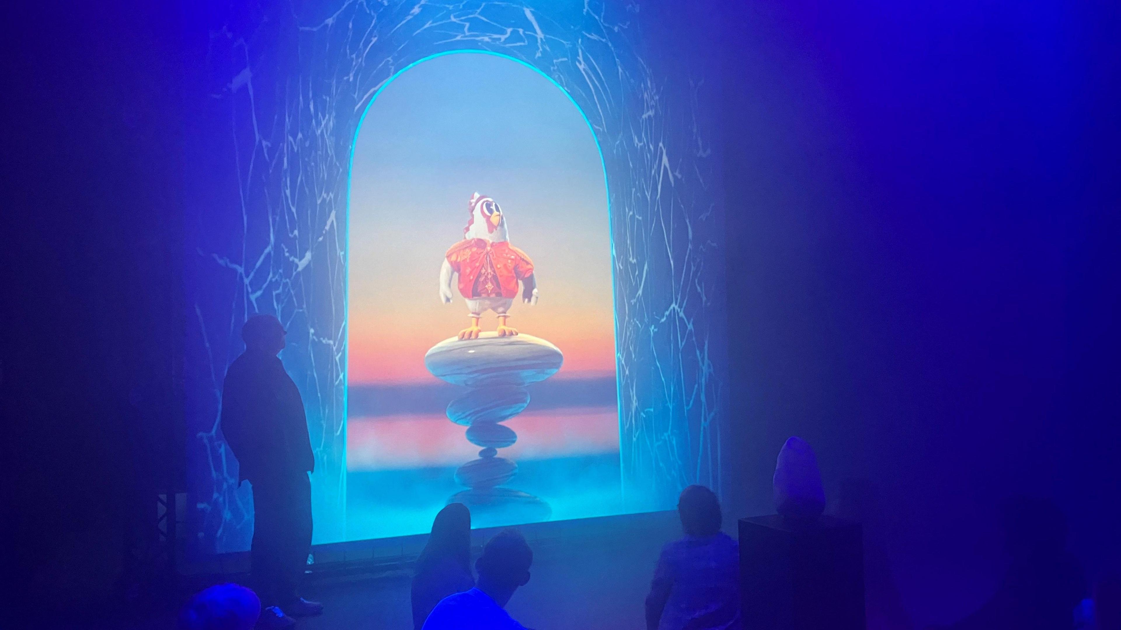 A cartoon chicken standing on a pedestal projected onto a screen in a darkened, blue room