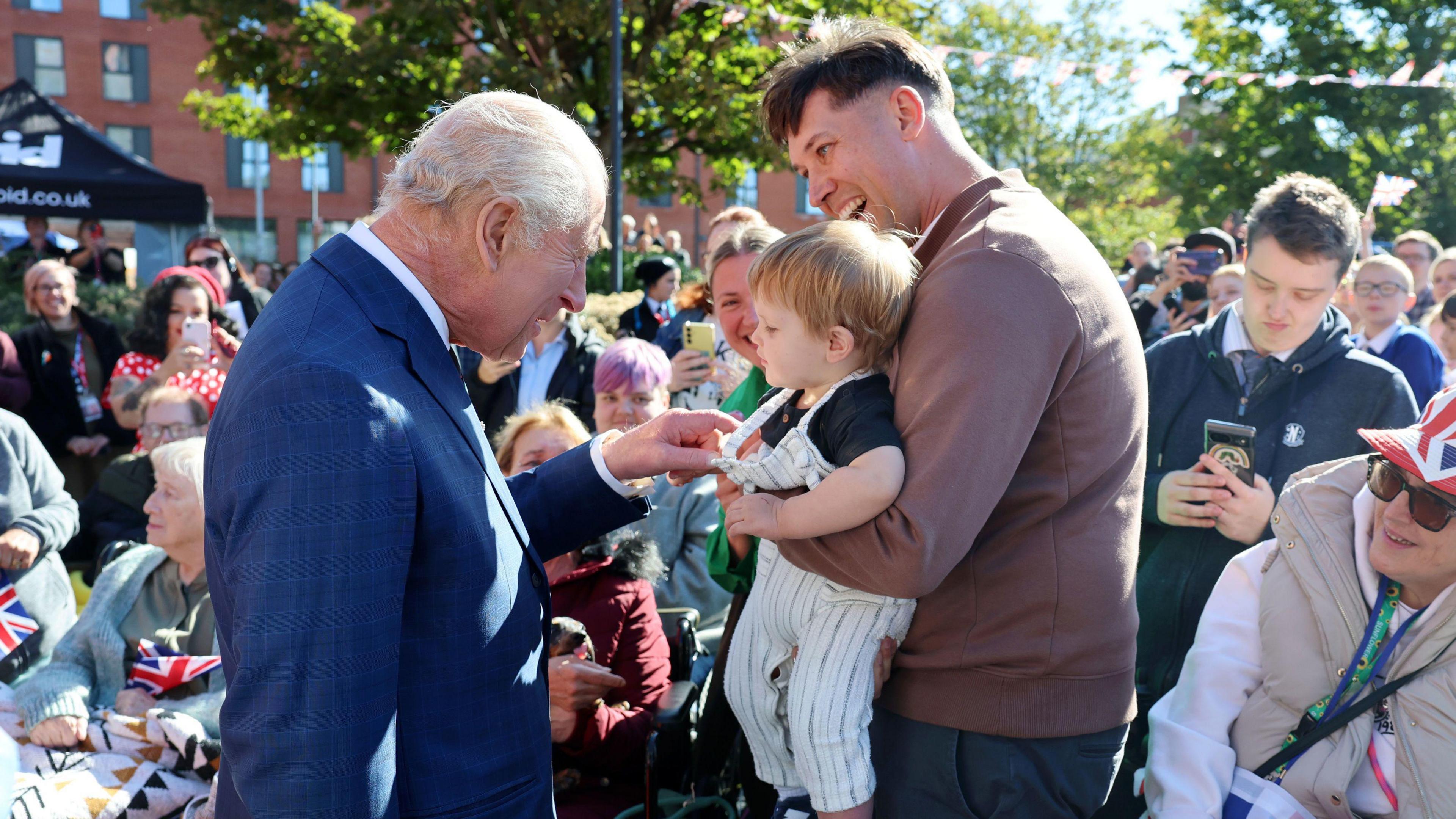King Charles III, on the left-hand side of the image, smiles as he greets a young boy being held up by a man.
