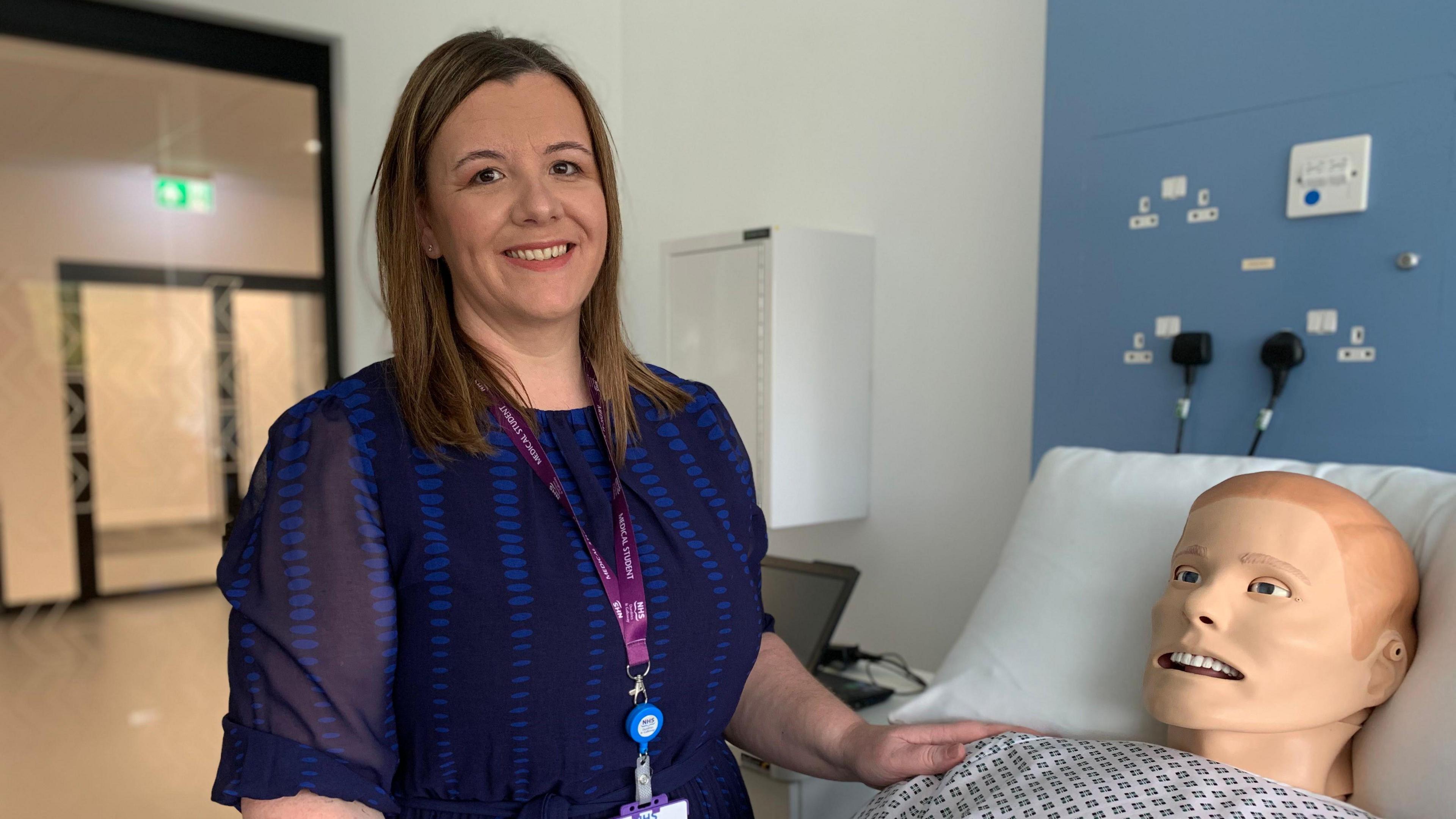 A woman in a blue dress with a purple lanyard round her neck with long, brown hair stands next to a model of