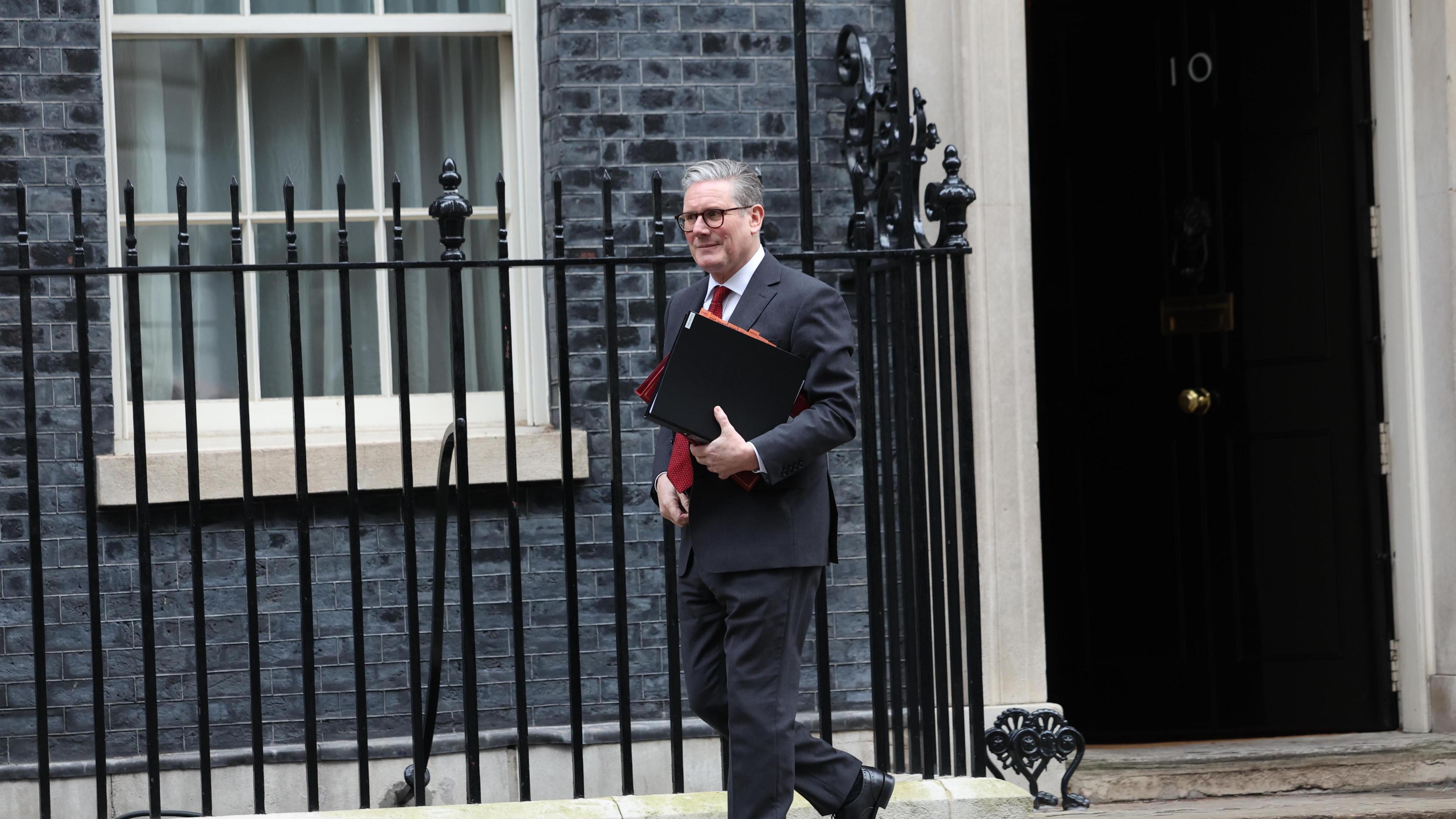 Prime Minister Sir Keir Starmer leaves 10 Downing Street. The black door with 10 on it can be seen in the background as Sir Keir walks away. He is wearing a dark suit and glasses and is holding a binder. 