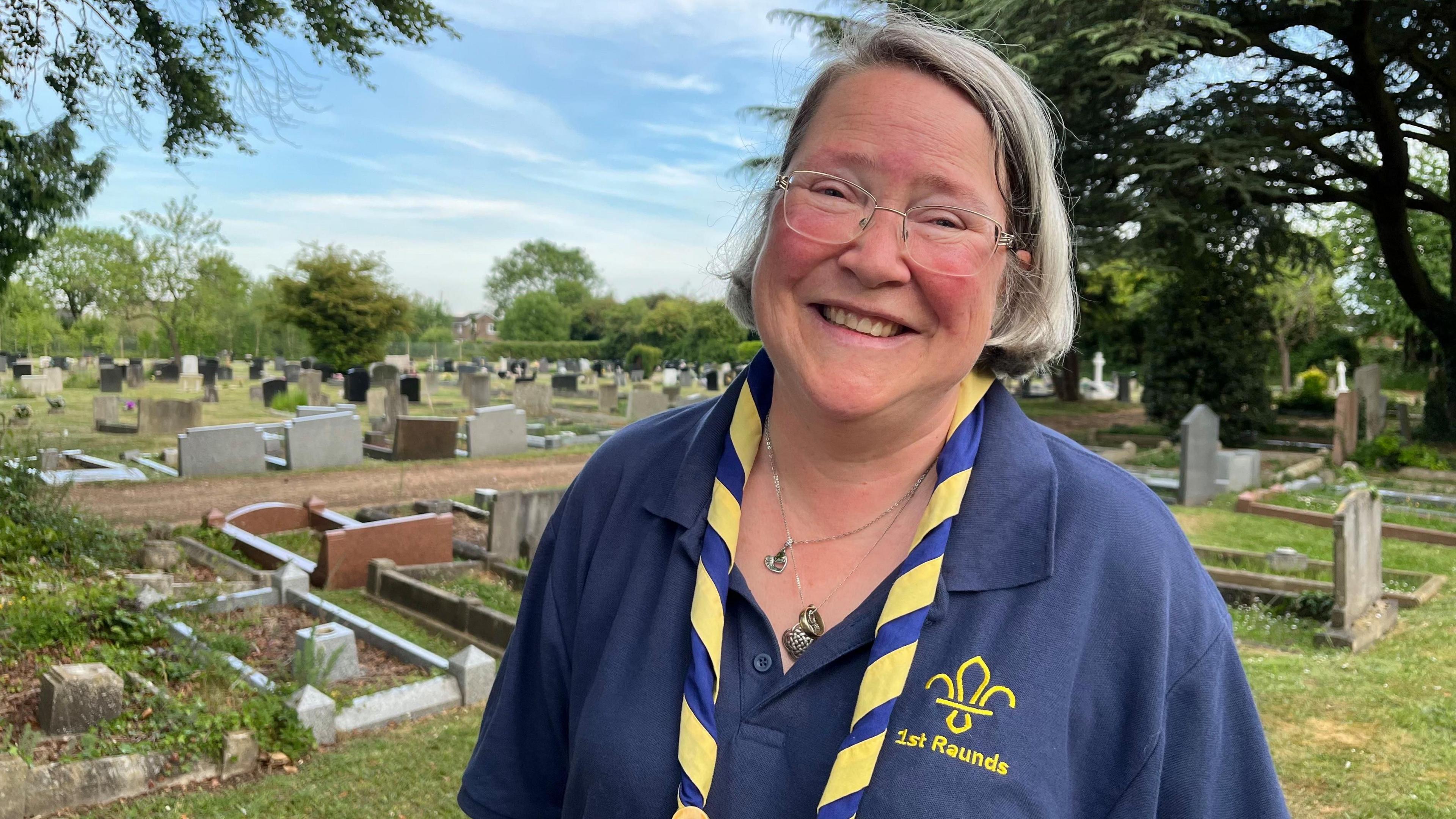 Woman with blonde hair and glasses standing in front of graves, with a blue polo shirt on. She has a blue and yellow ribbon around her neck.