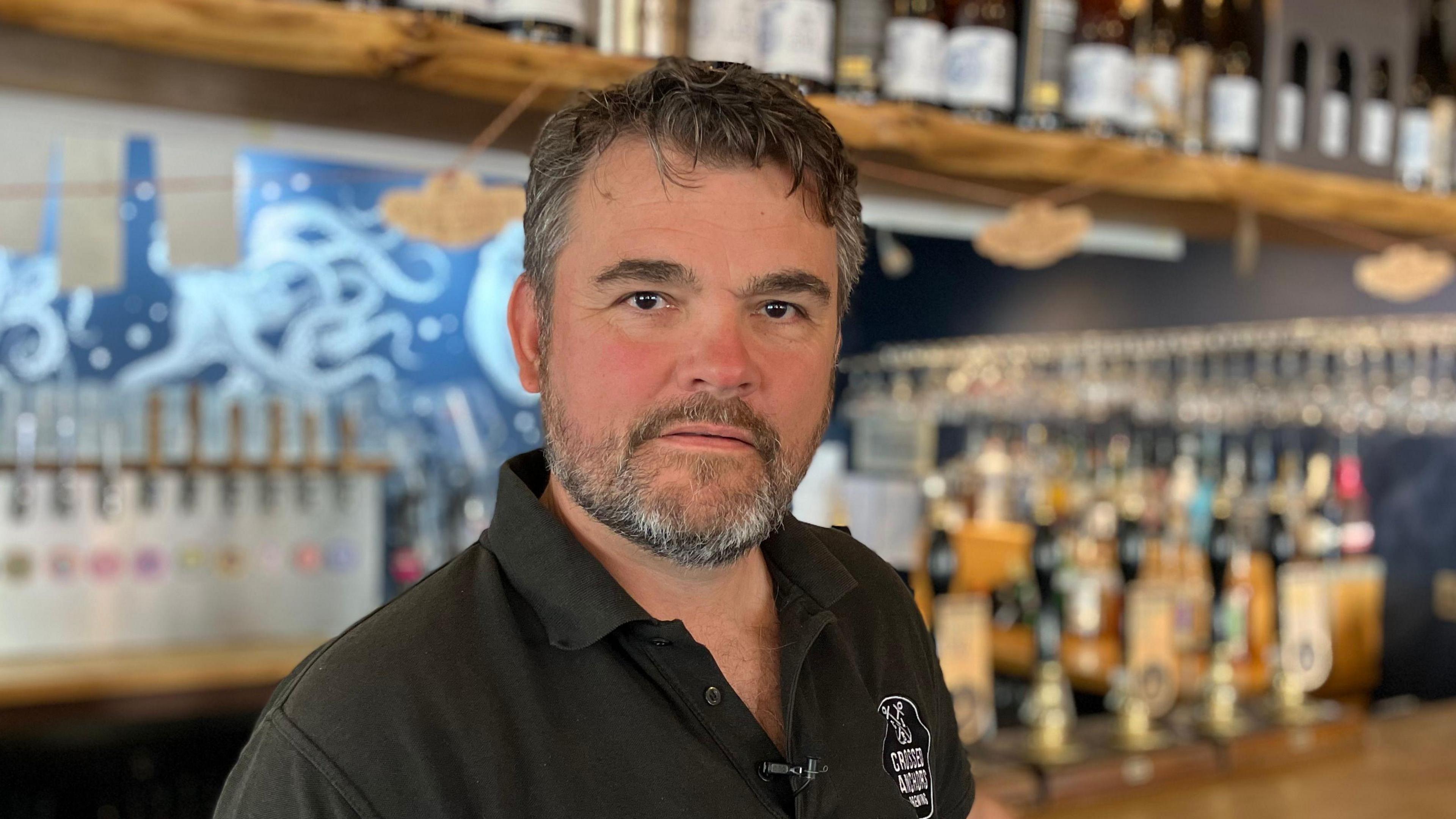 Oliver Bainbridge wearing a black polo shirt stood in front of a bar he owns.