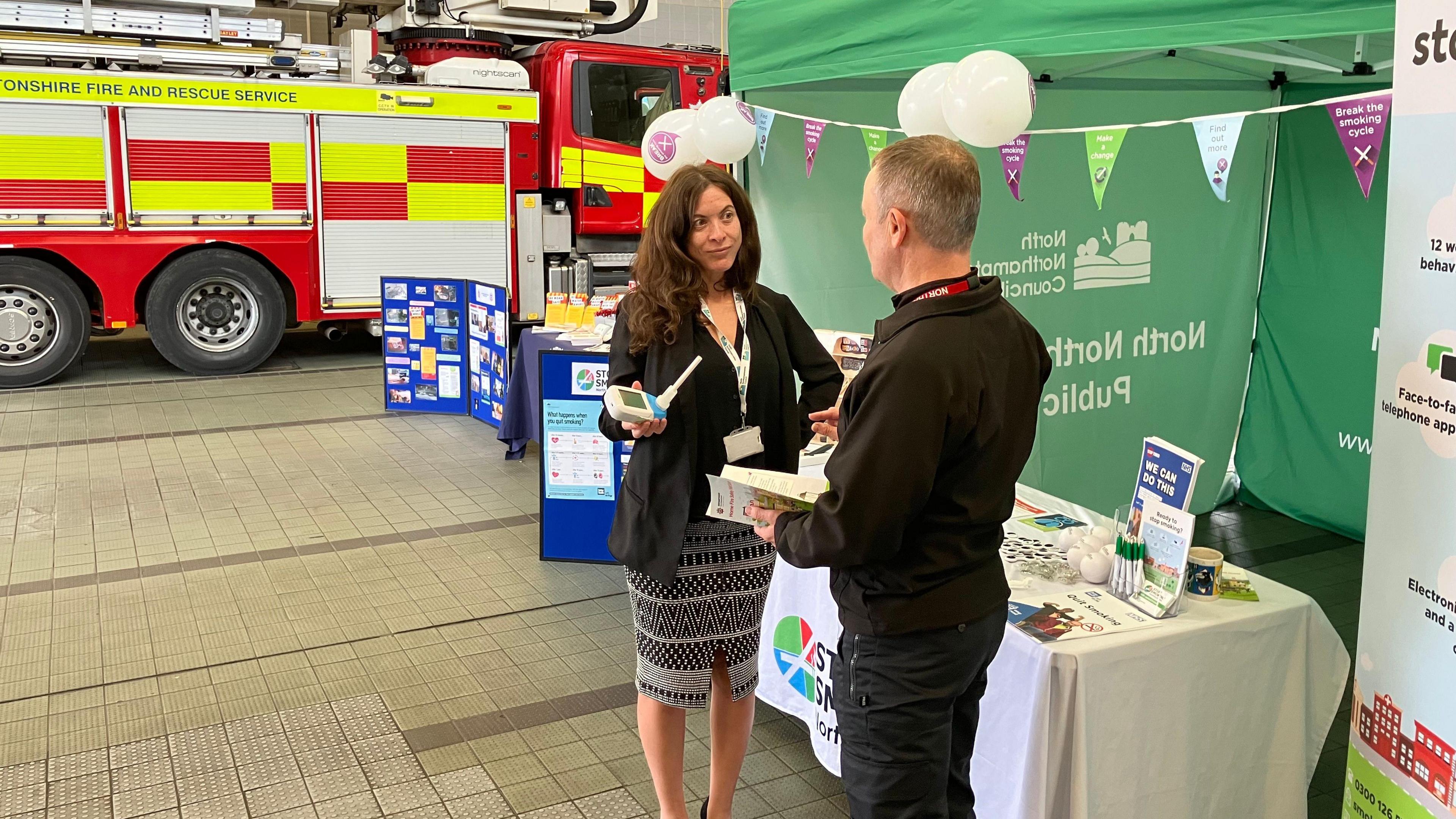 Two adults are standing in front of a stall that has been set up inside a fire station. There is a green gazebo with a white table in front of it. The table has leaflets laid out on it. There is colourful bunting and white balloons on the gazebo. A woman and man are talking while holding a small piece of white equipment and a leaflet. In the background is a fire engine.