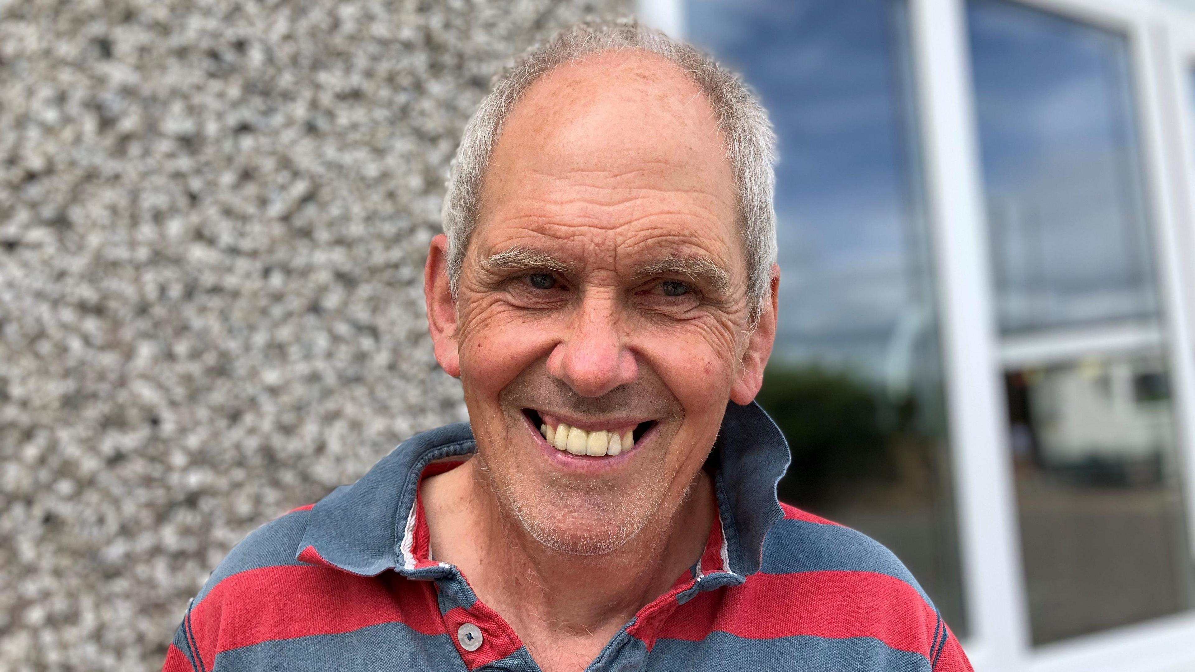 Older man with grey hair smiling at the camera. He is wearing a red and grey striped polo shirt 