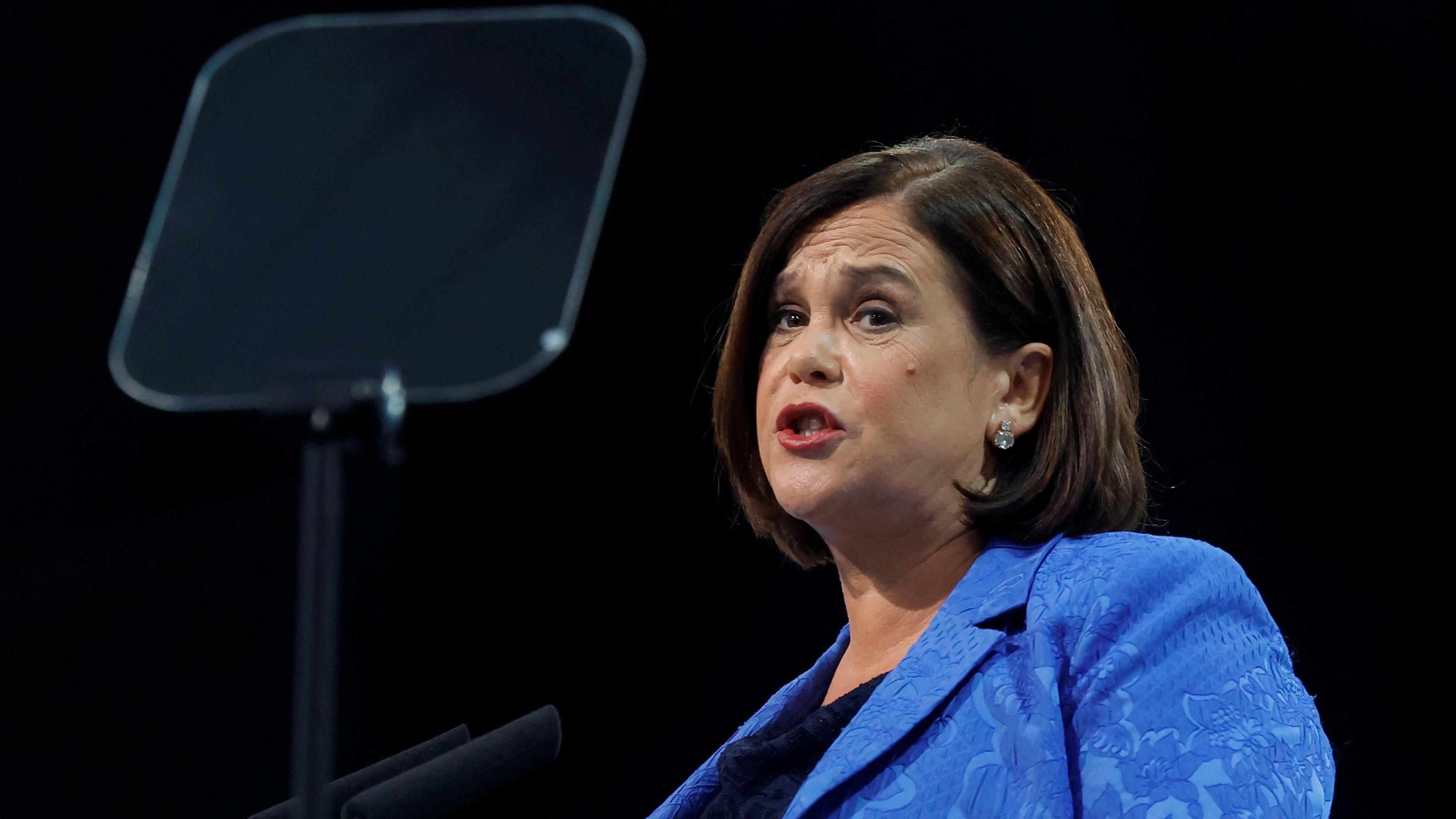 Mary Lou McDonald speaking at a microphone against a black background. In the left of the shot is a glass teleprompter 