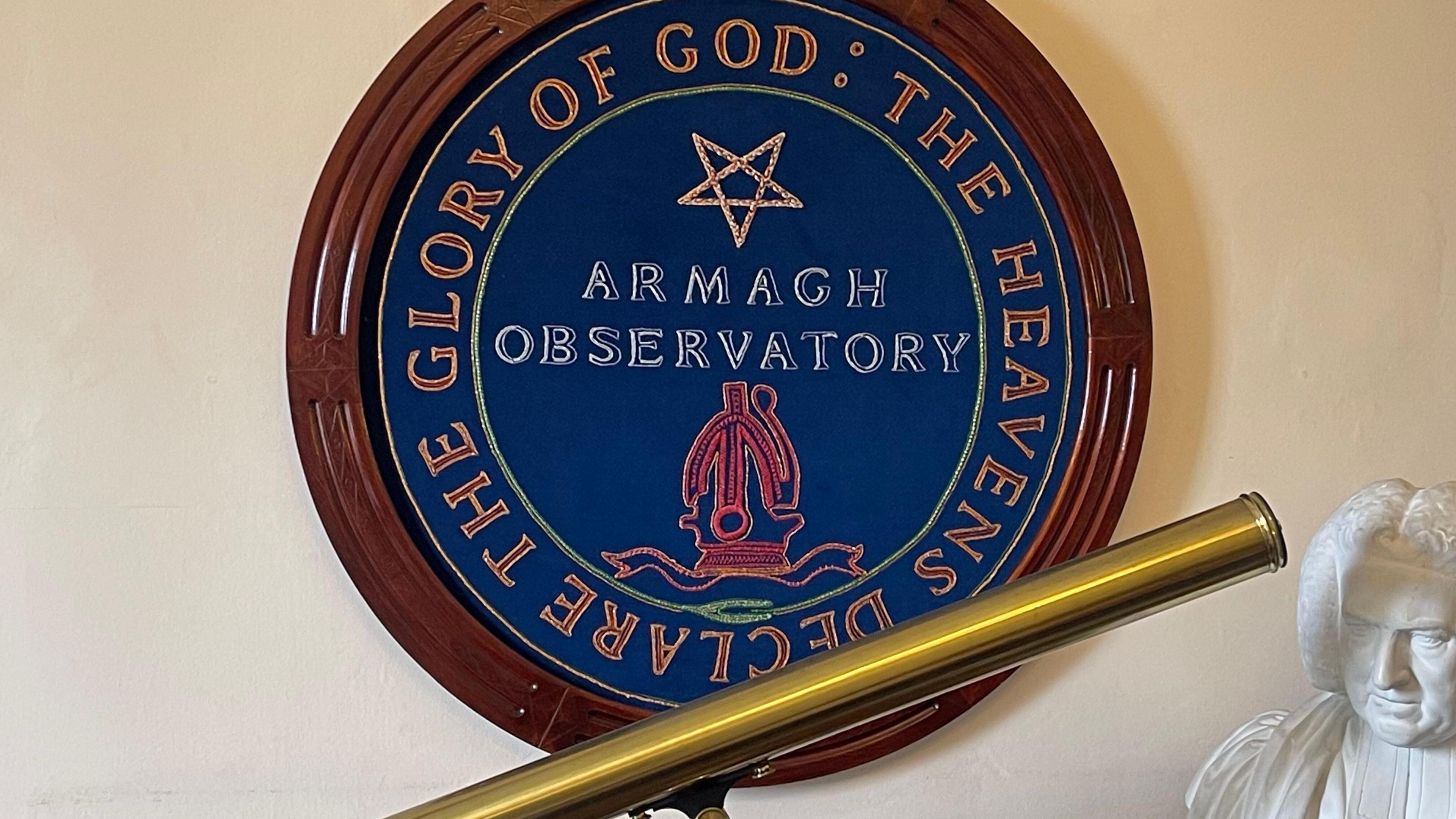 A large Armagh Observatory crest with a wooden frame on a wall. A shiny brass telescope on a shelf under the crest, along with a with bust of a Georgian era man.