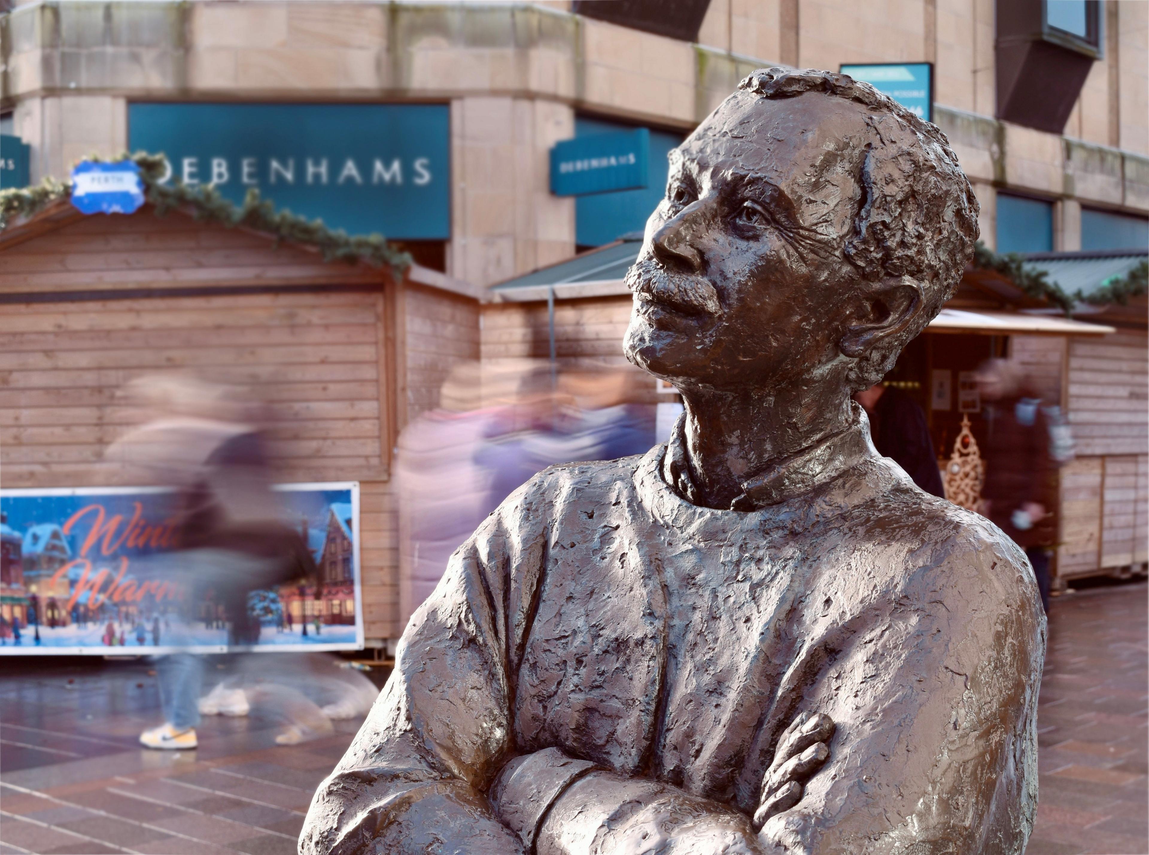 Bronze sculpture of a man with his arms crossed, located on a busy street as blurred shoppers rush by in the background