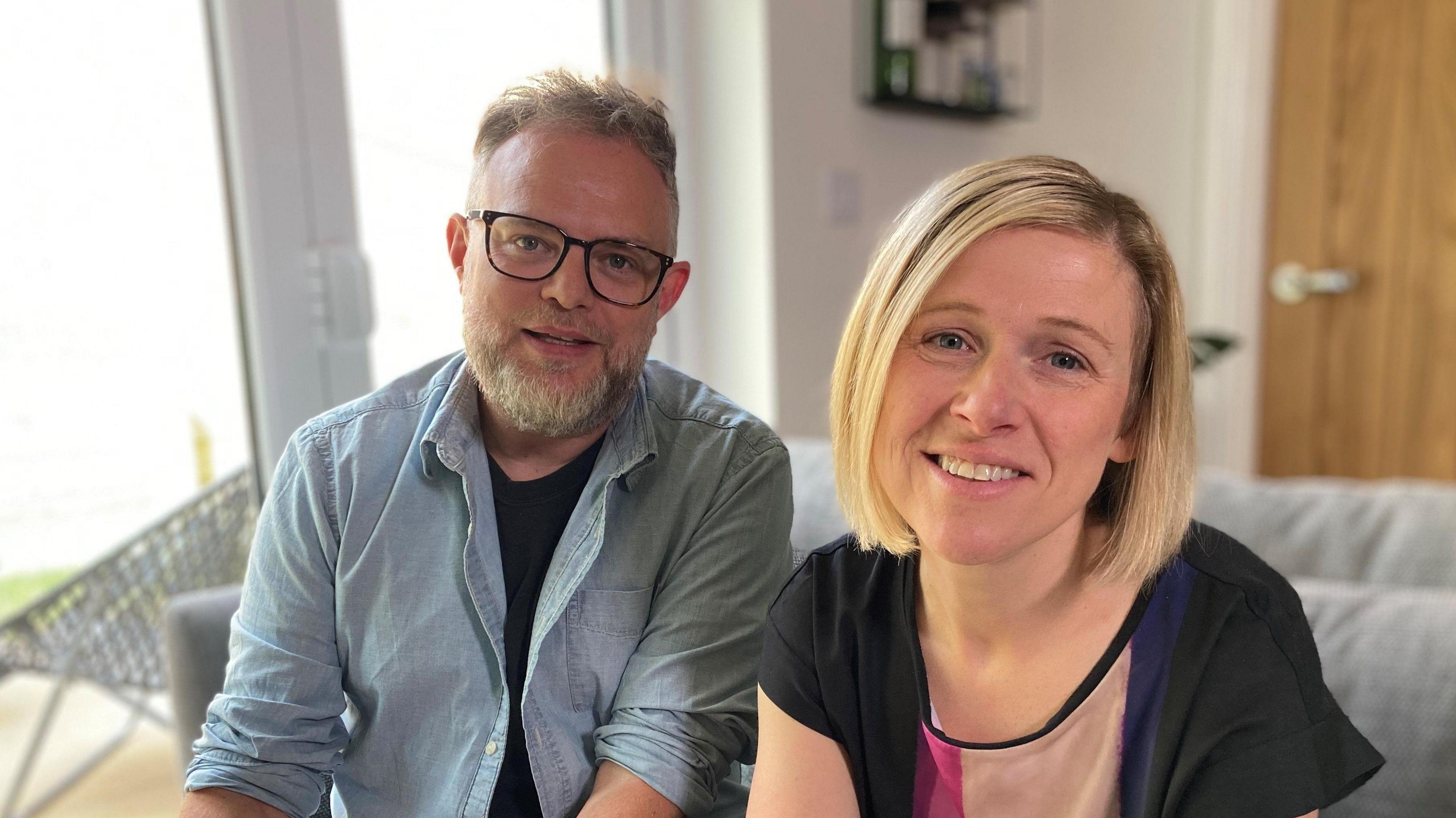 Macaulay's parents Ben and Rebecca are sitting on the sofa looking at the camera, Ben, on the left, has short grey hair and a beard and is wearing black glasses. Rebecca has mid-length blonde hair.