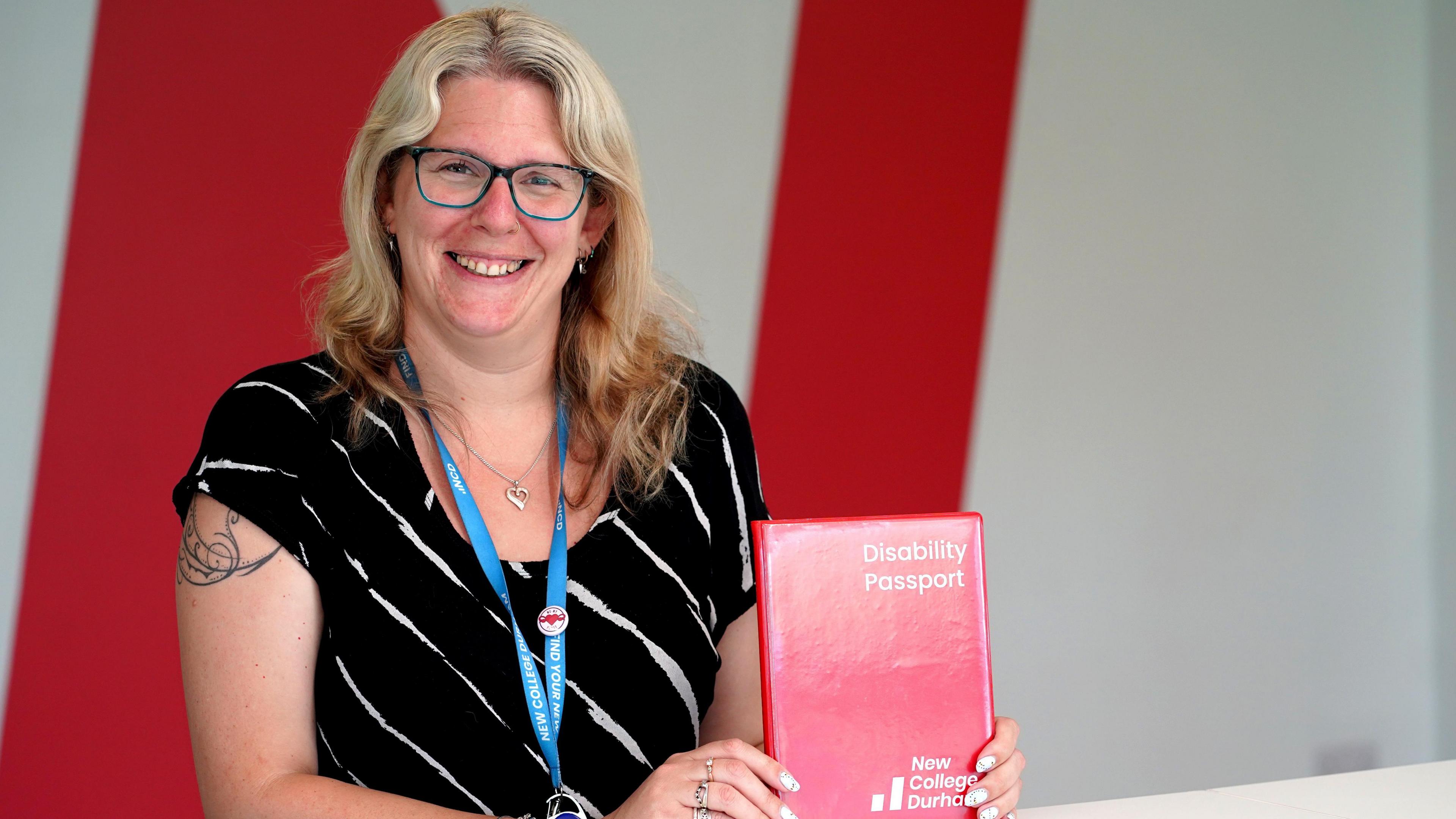 Mhairi Vale is smiling as she holds the disability passport. It is a small book with red covers reading Disability Passport New College Durham. Mhairi has blonde wavy hair and is wearing a black t-shirt and glasses.