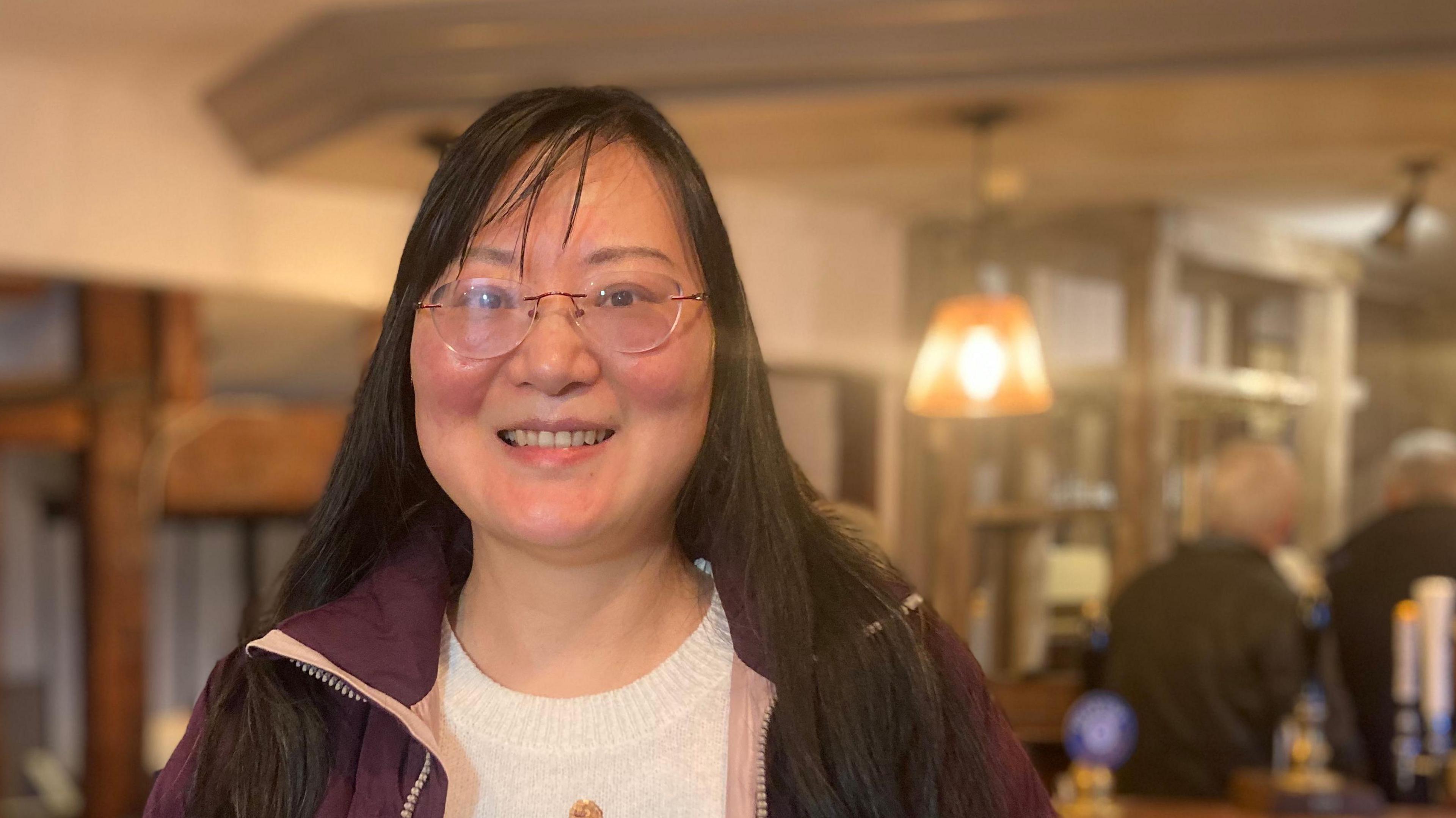 Chulyal White is smiling at the camera. She has long, dark straight hair and is wearing spectacles and a maroon jacket. People can be seen at the bar in pub behind her