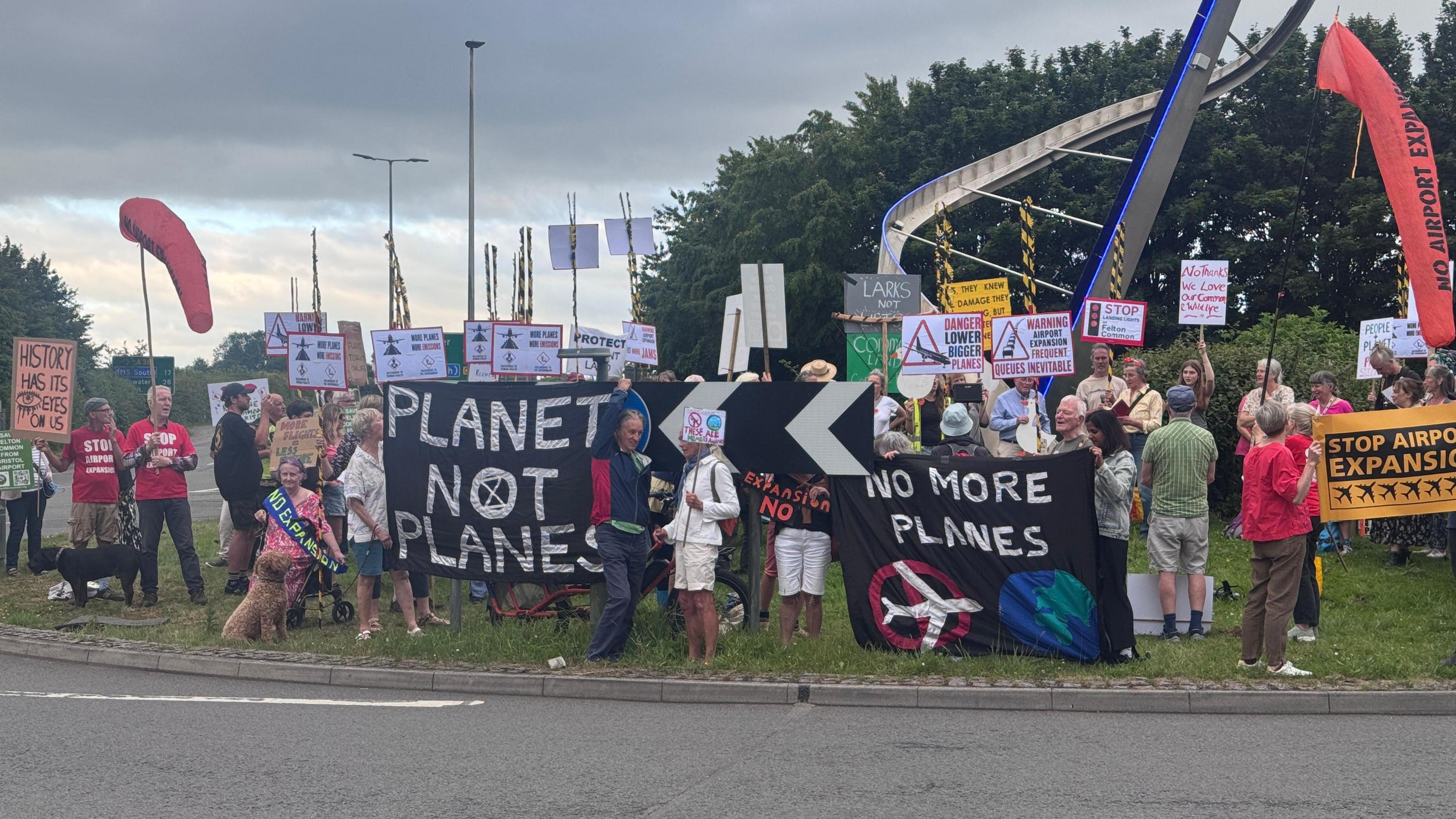 Protesters by the side of a road at Bristol Airport hold up banners and flags, which read 'People No Planes' and 'No More Planes'.