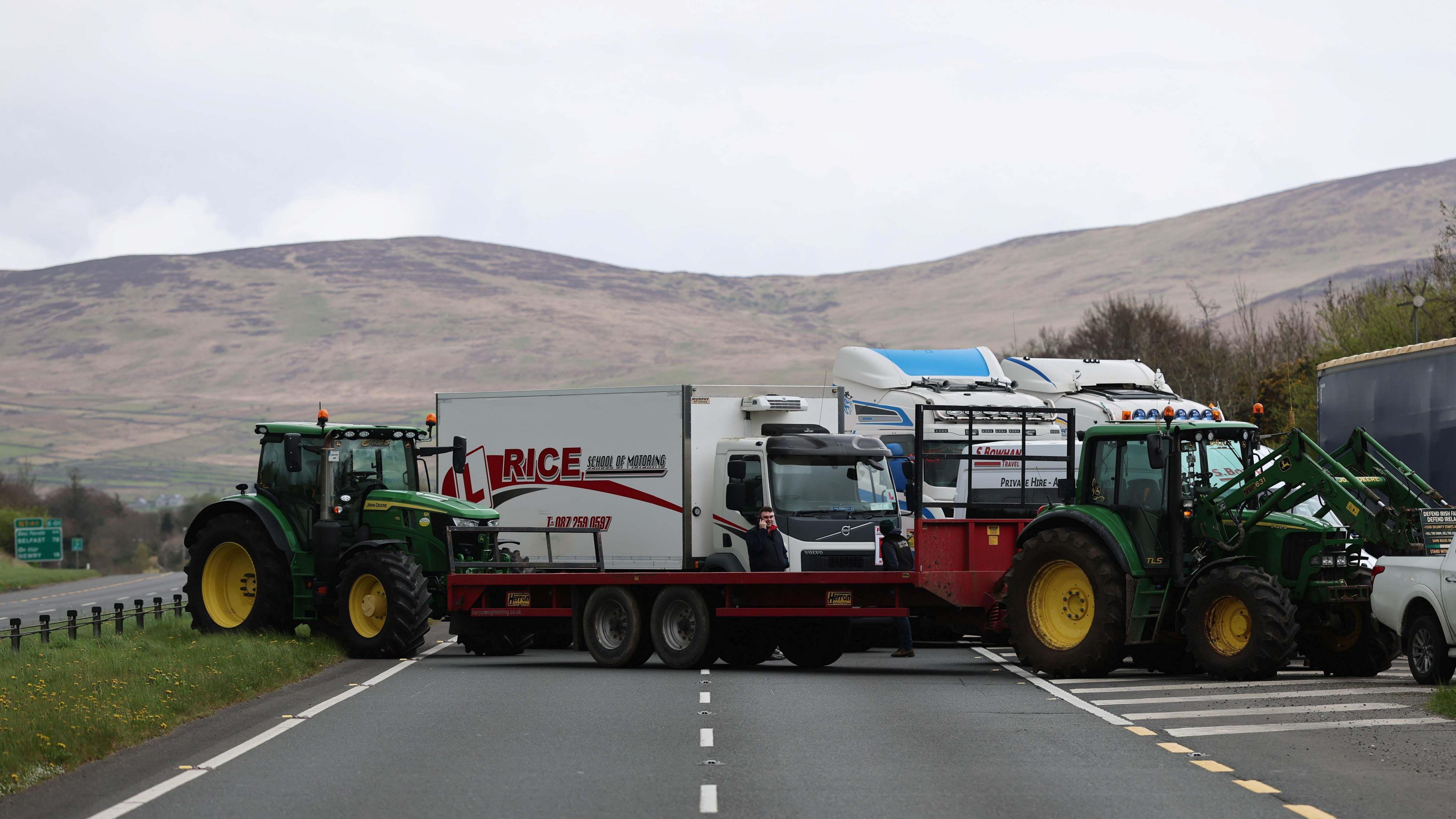A road blocked by a number of tractors and trucks which are parked in the middle. In the background there is a green sign which has 'BELFAST' and 'NEWRY' written on it. 