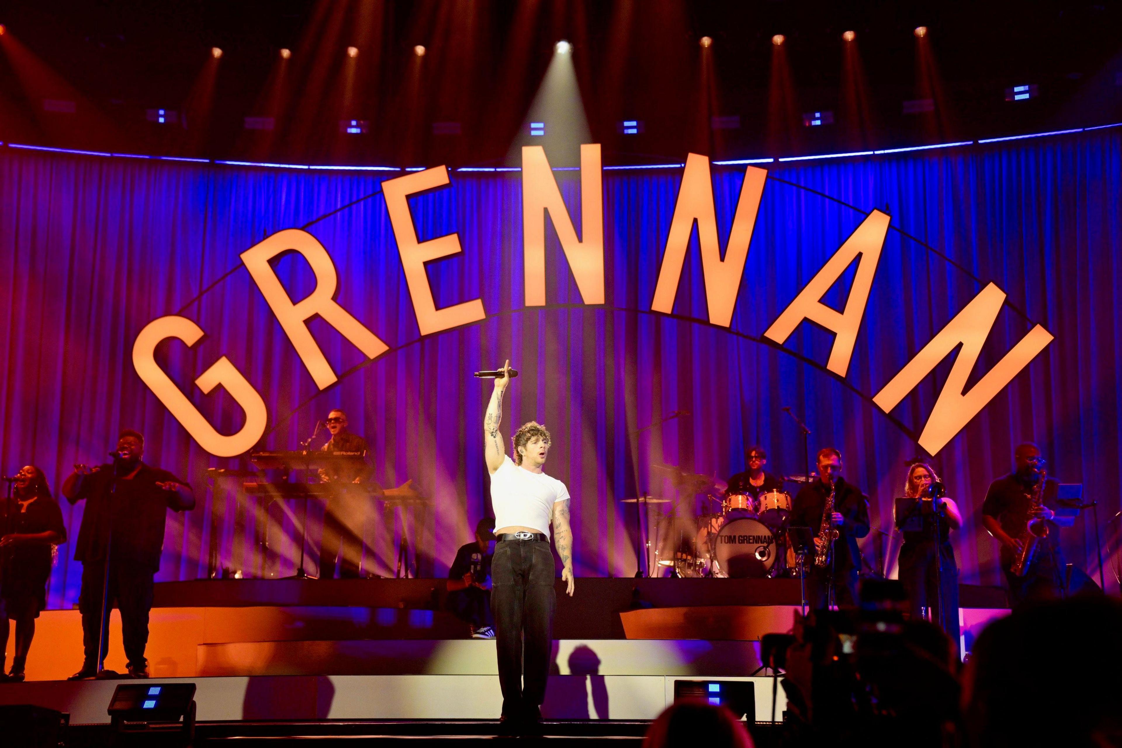 Singer Tom Grennan is on stage in front of a band. He is wearing a white T-shirt and black trousers. He has one arm raised in the air. There are musicians and singers behind him and he word "Grennan" is in large letters above the stage. 