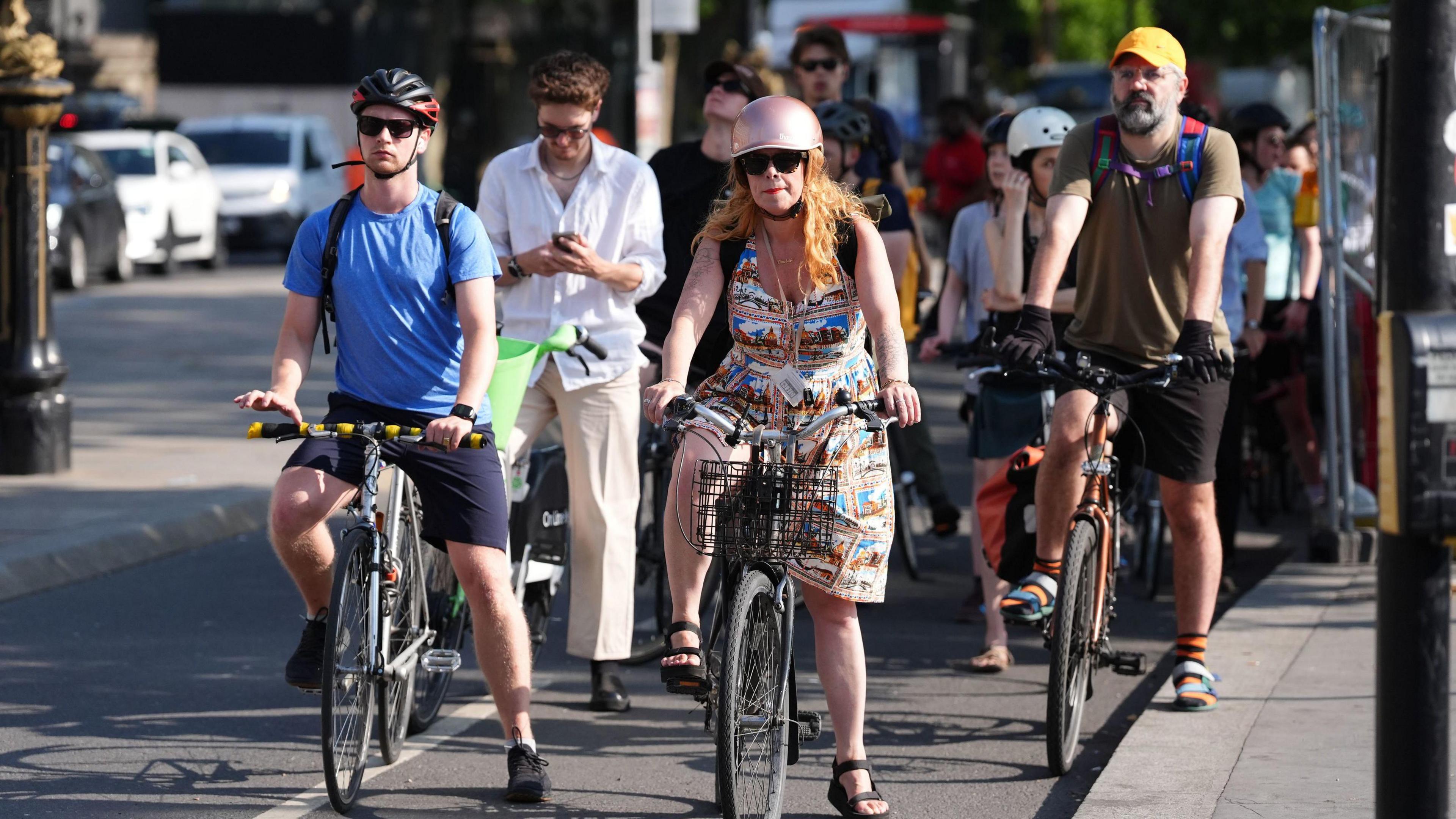 Cyclists pause on the road, some wearing helmets, as the sun shines down.