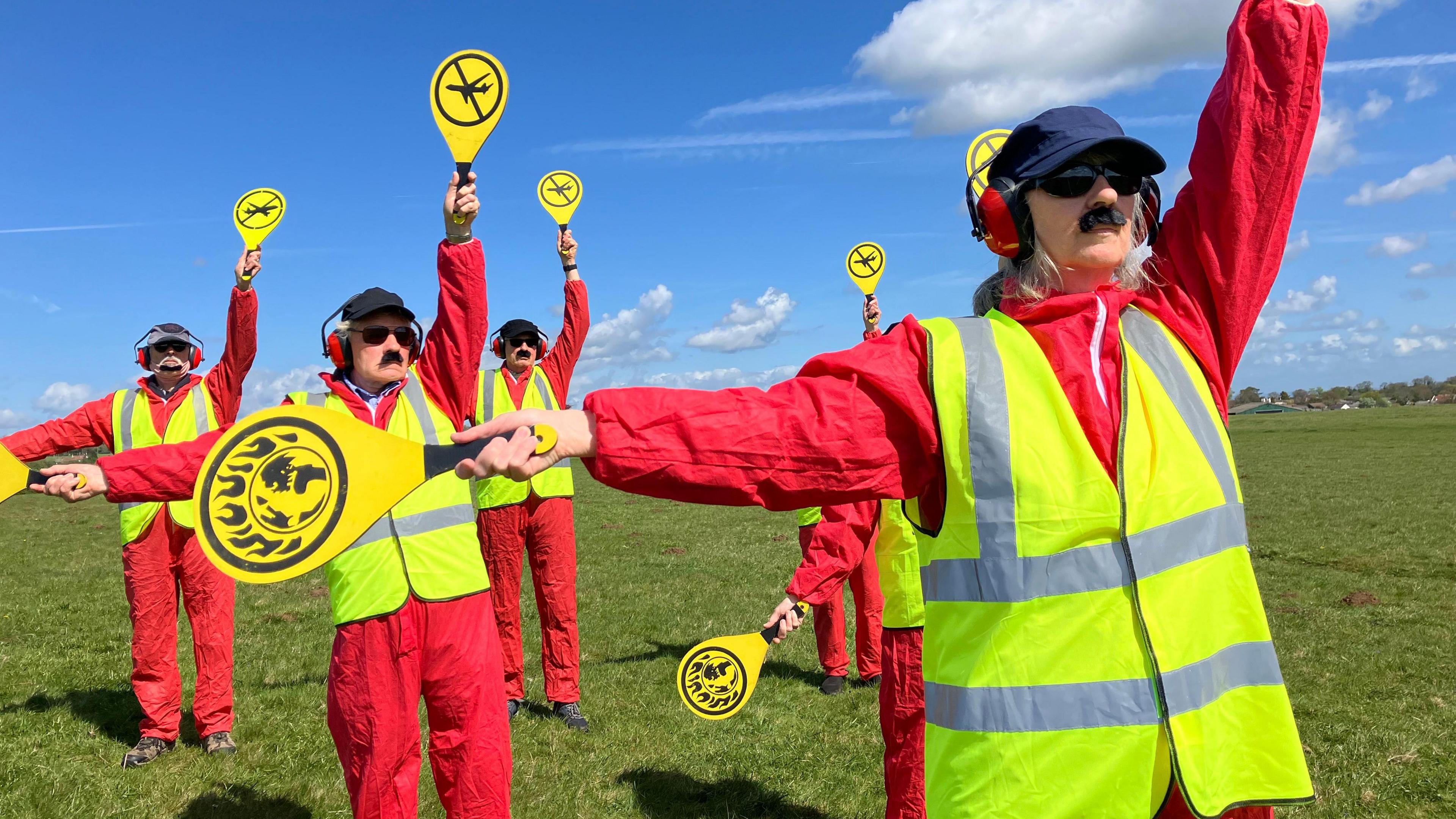 A group of people are dressed in red jumpsuits with yellow high-vis vests holding up yellow signals.