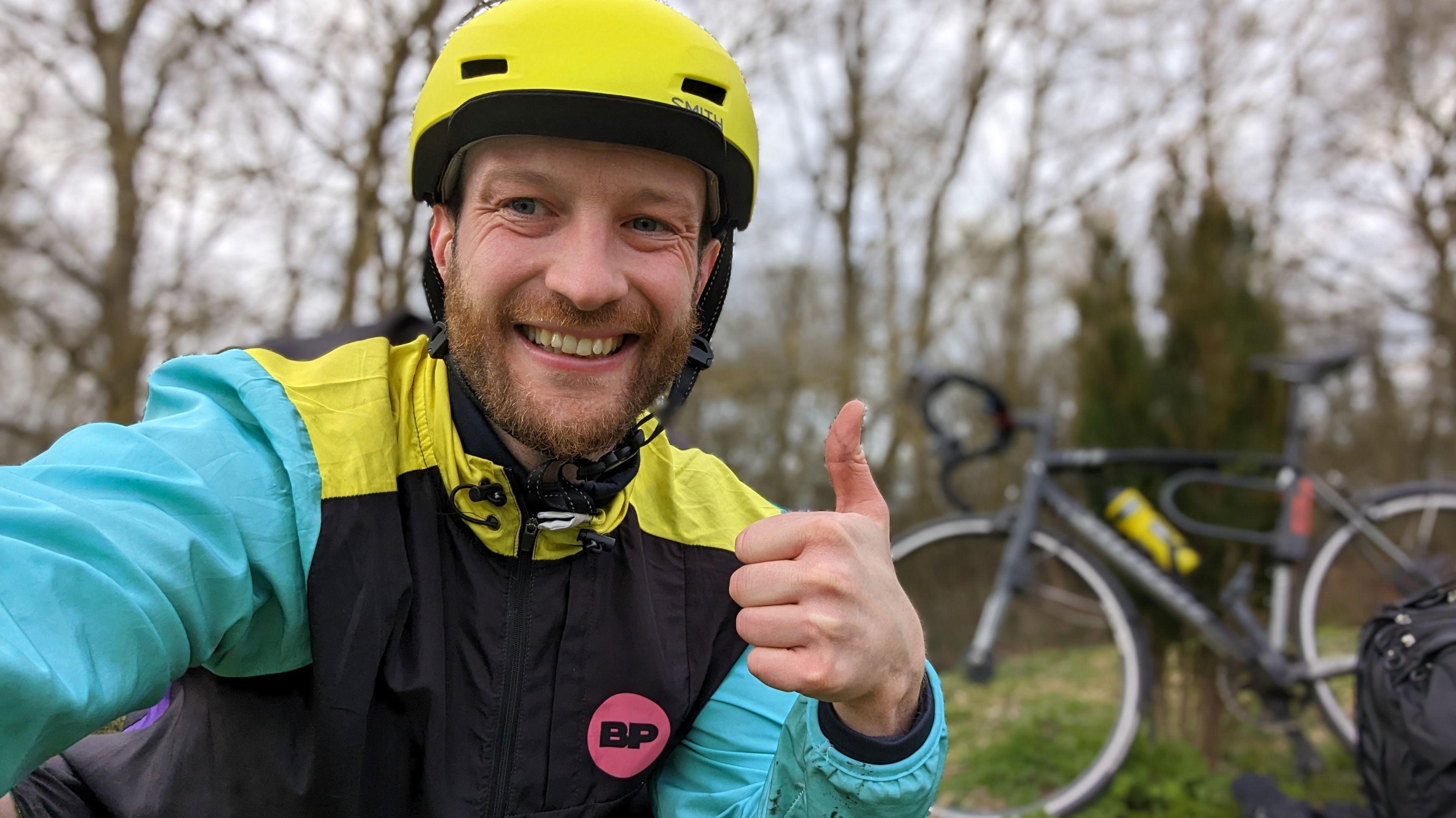 James is crouching in front of the camera and has his thumbs up. He is wearing a bright yellow bike helmet and a multicoloured waterproof jacket. 
In the background, out of focus you can see his black bike. 