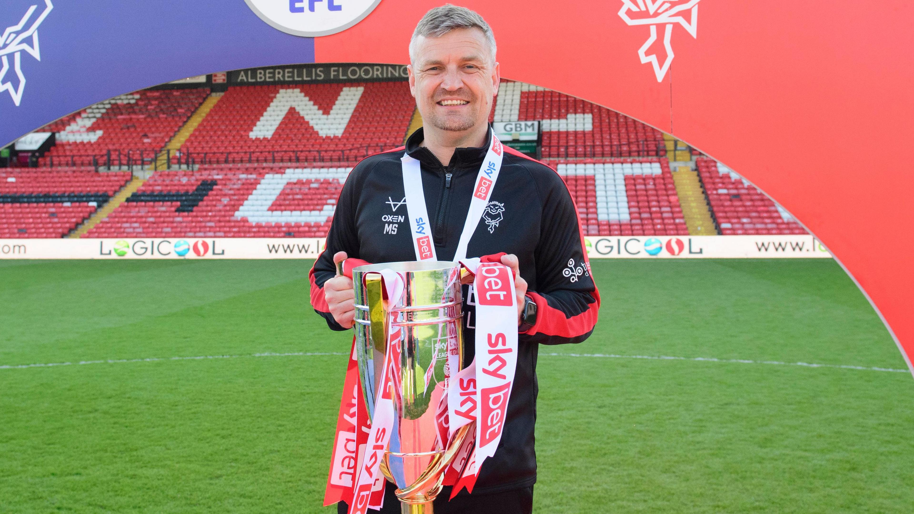 Michael Skubala, wearing a black Lincoln City tracksuit top, holds the League One trophy in his hands. The red seats of Lincoln's LNER Stadium can be seen behind.