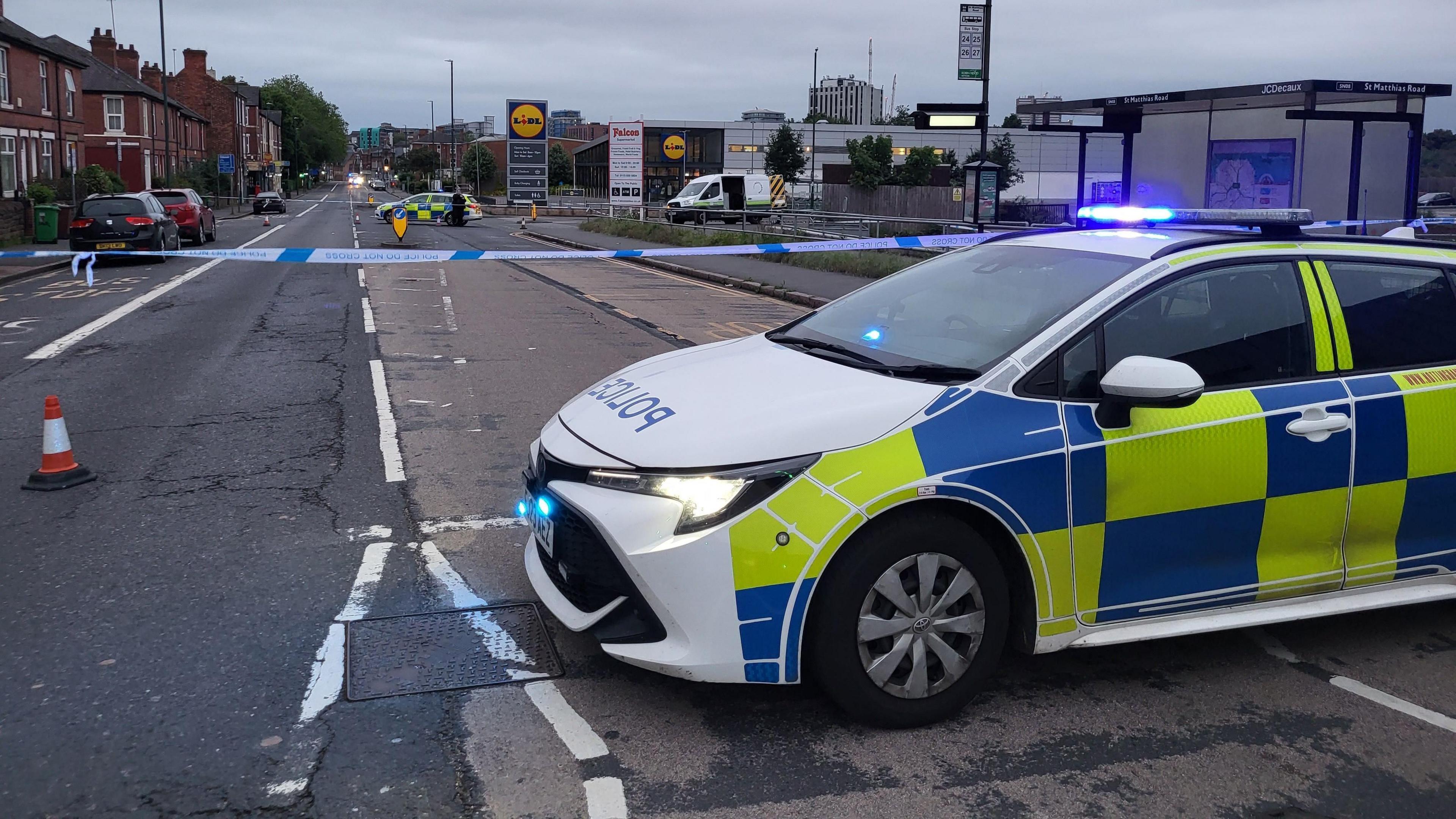 A police car is parked in front of police tape stretching from one side of the road to another. Another police car can be seen in the cordon.