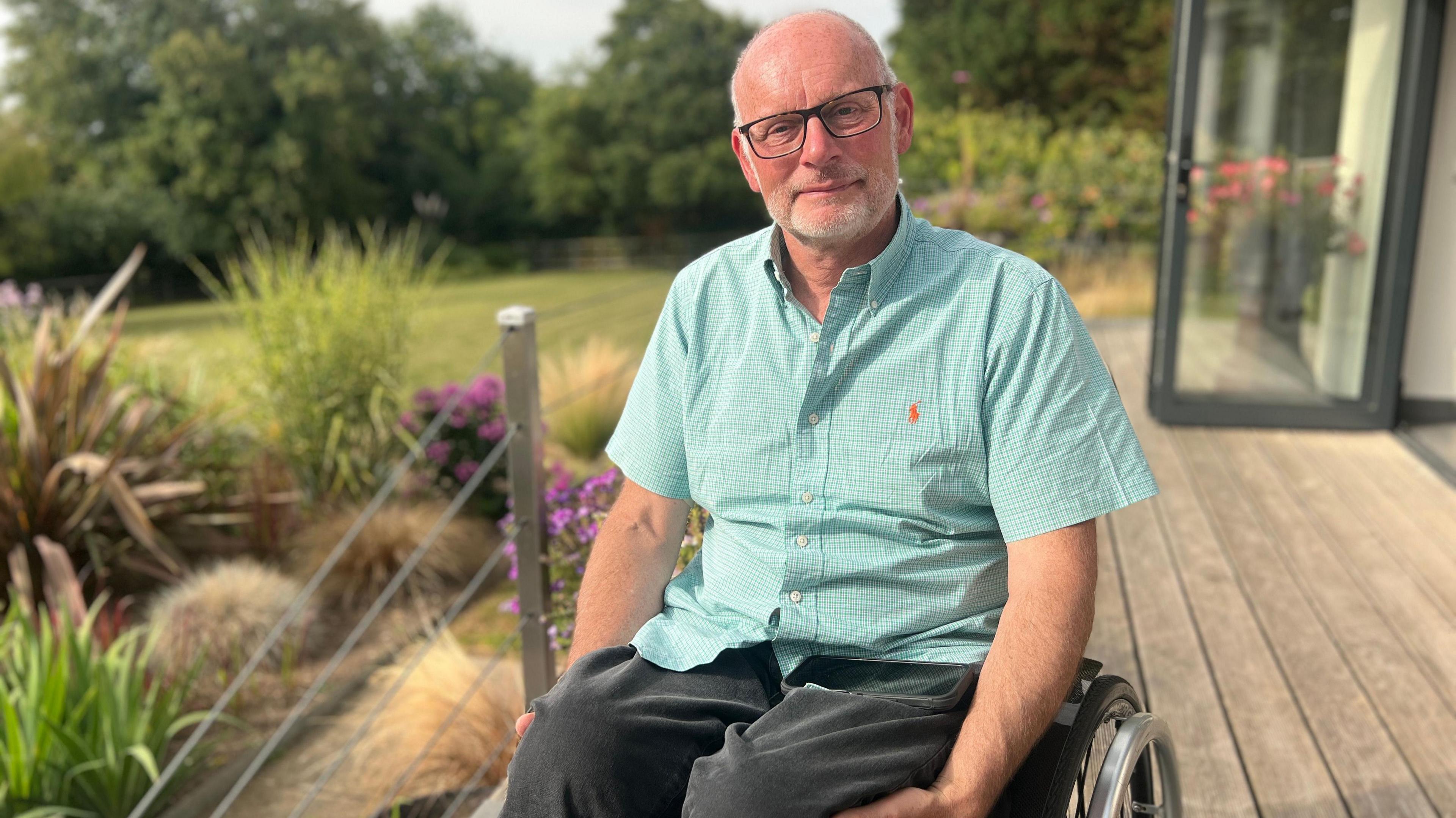 Keith Carter looking into camera on his decking overlooking his back garden. Keith has dark rimmed glasses, has short white hair and stubble. He is a wheelchair user and is wearing a turquoise shirt and dark trousers.