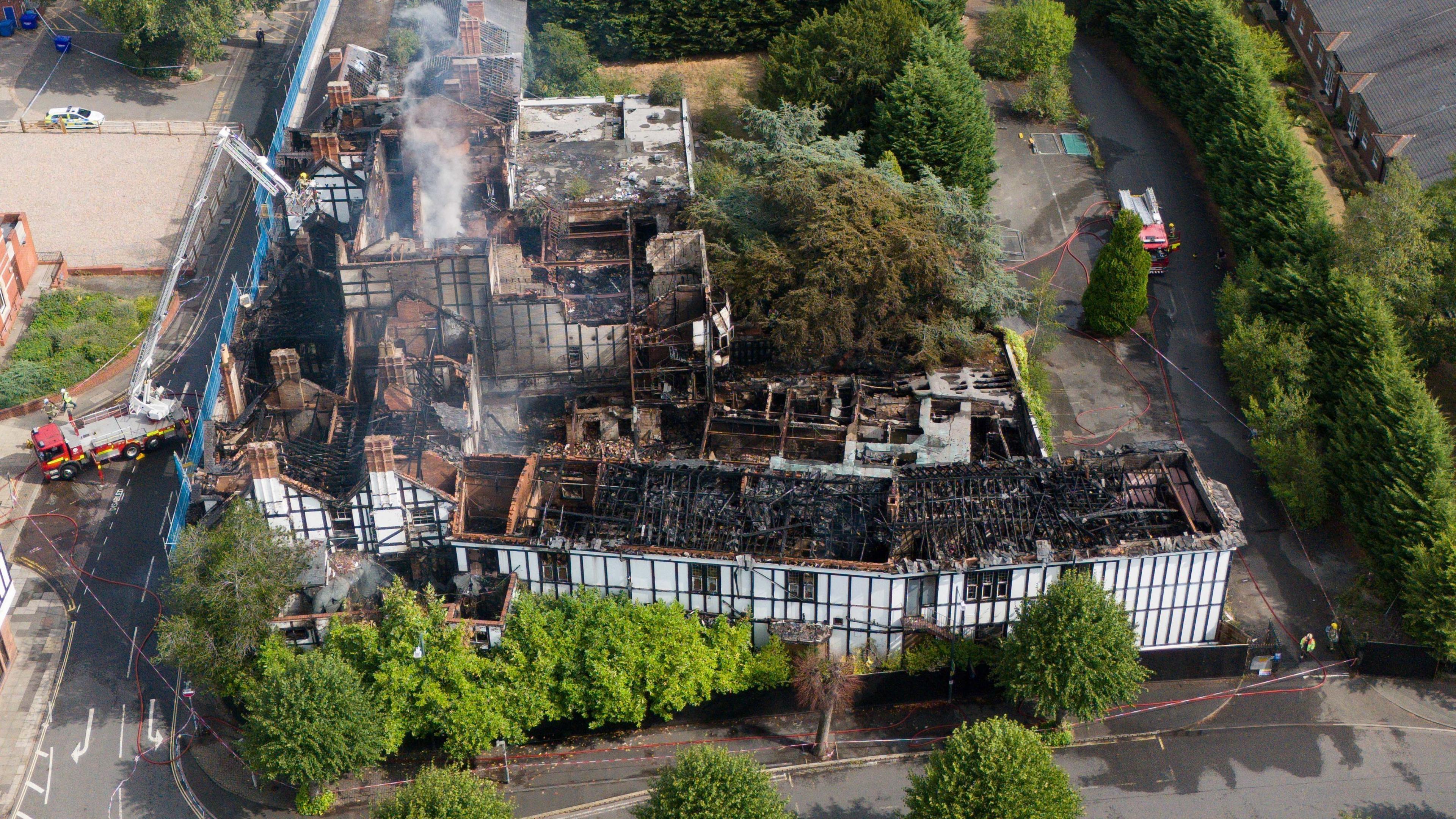 An aerial drone photo of the raven hotel. It is a black and white building and the roof has been destroyed by fire. It is open and reveal blackened beans and insides, with grey smoke rising from it.