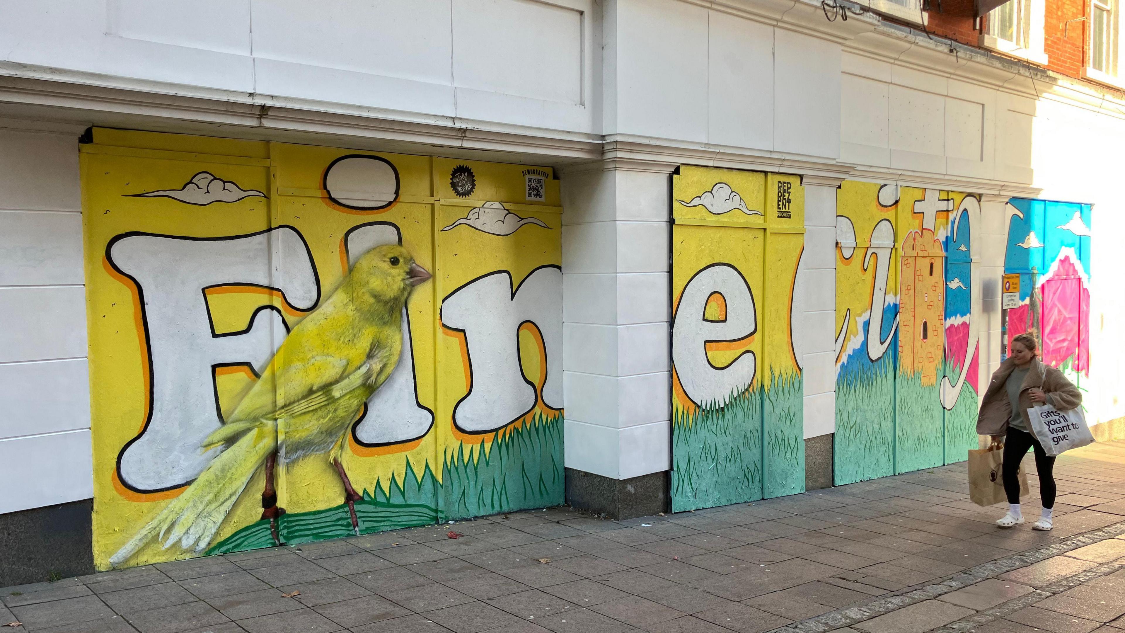 Mural reflecting Norwich's association with a canary. Large painting of the bird is on a building's wall. A shopper walks past the building. 
