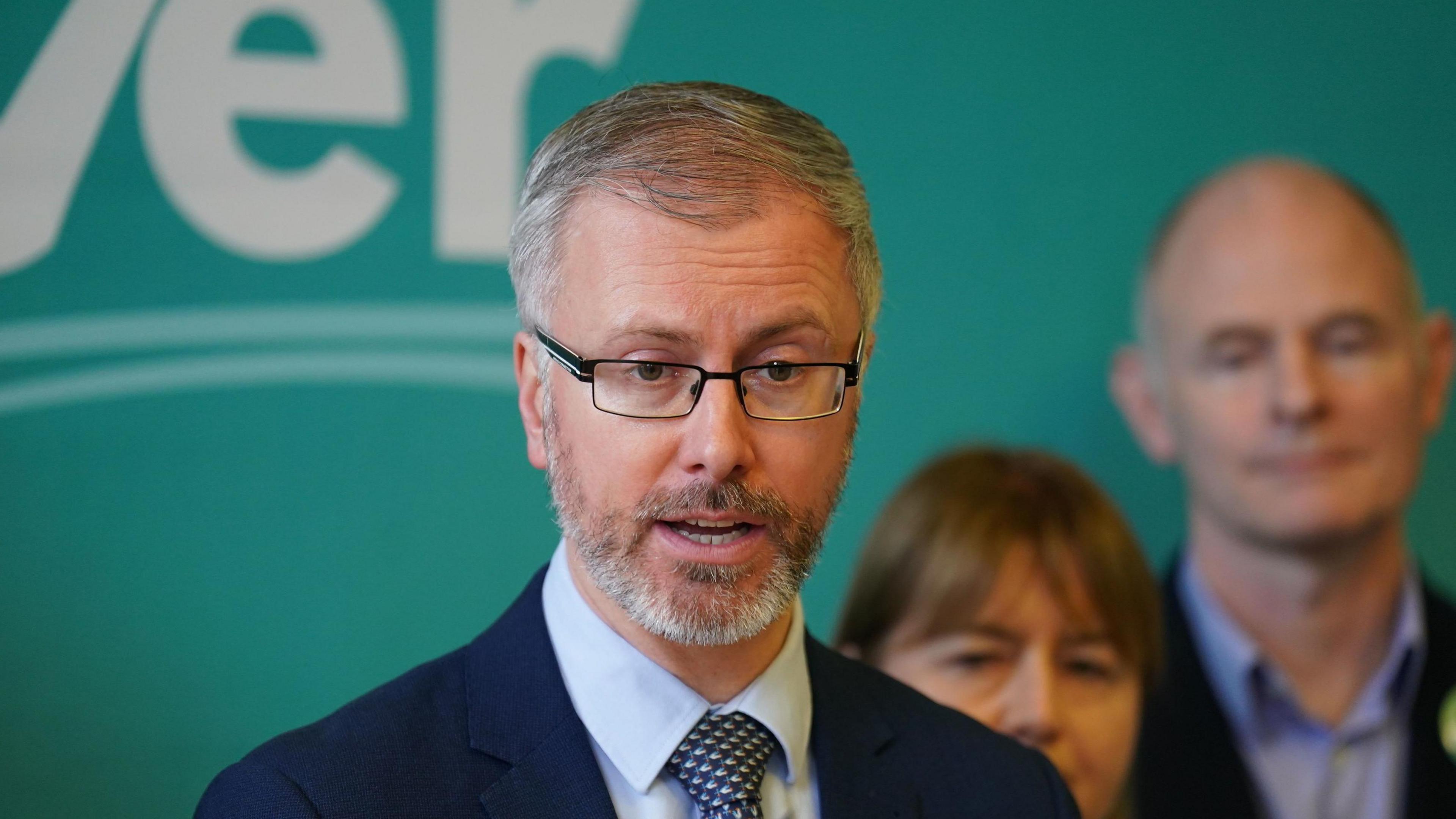 Green leader Roderic O'Gorman, with party candidates, speaking during a press conference at the Irish Architectural Archive, Dublin. He has greyish hair and brown eyes. He is wearing glasses, a dark blue blazer, light blue shirt and check tie.