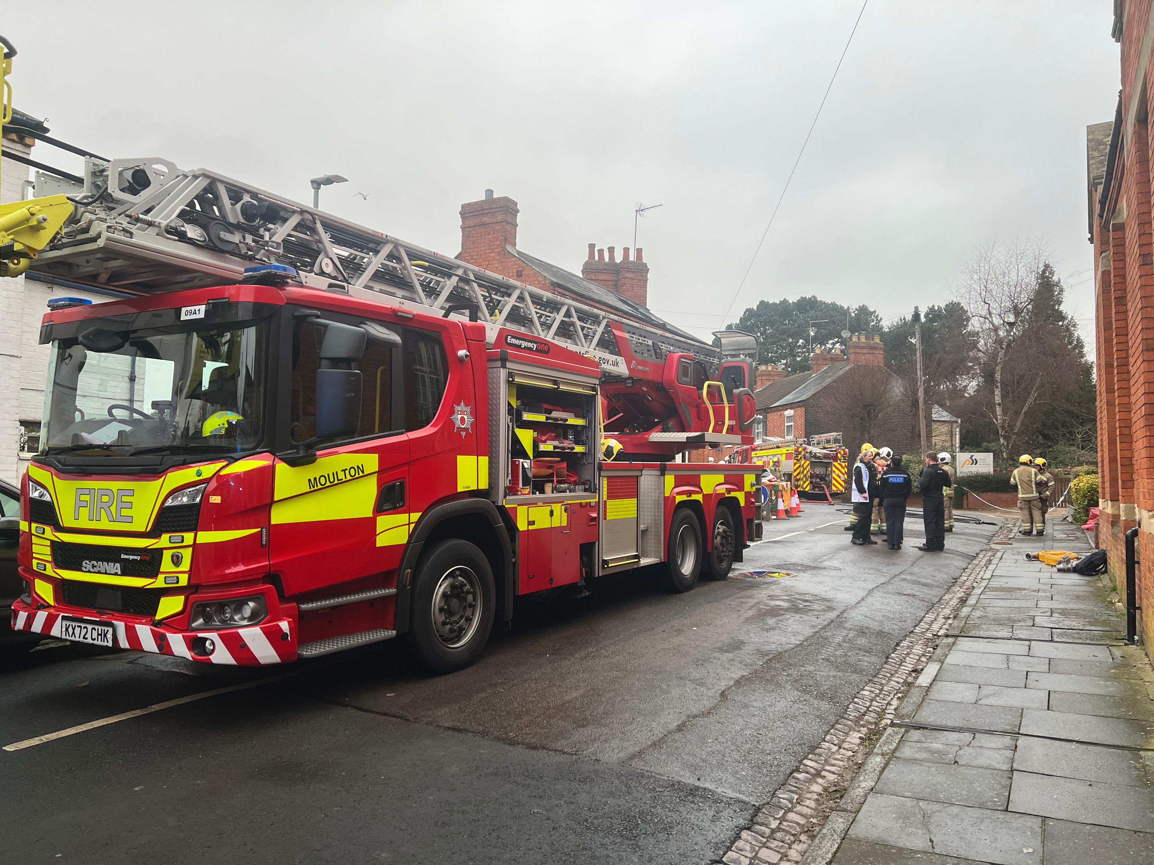 A fire engine with ladders parked in the middle of a road near a red brick building with a group of police and fire officers standing close to the vehicle.