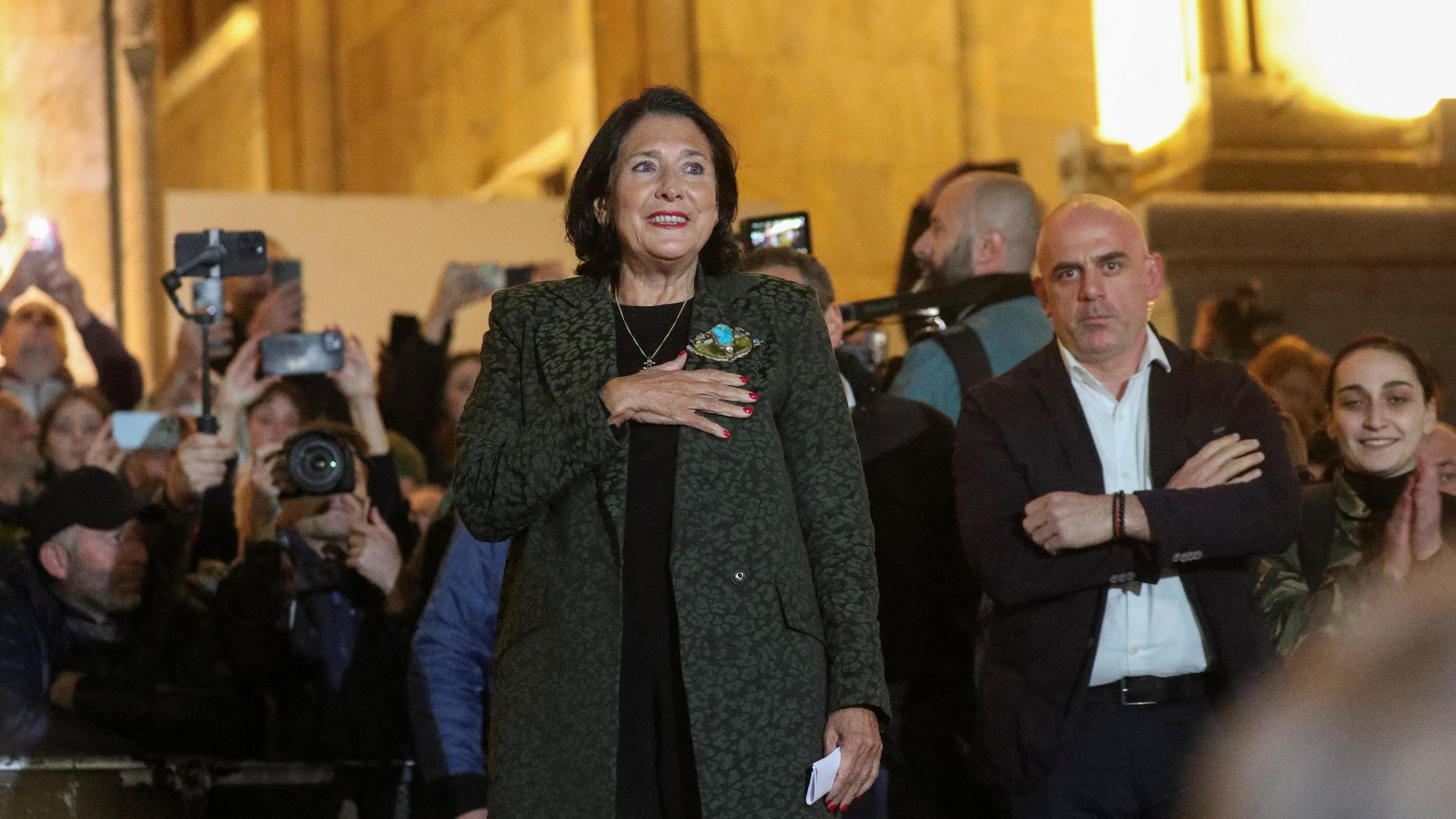 A woman in a grey coat standing with a hand on her heart. She is surrounded by people and photographers