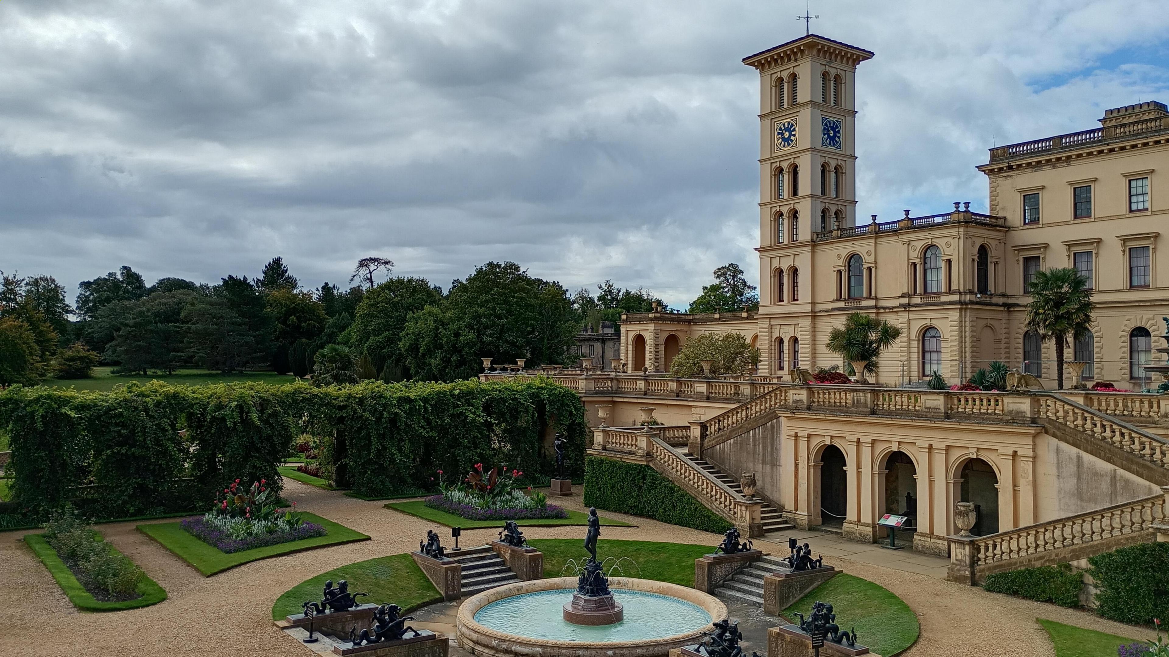 The stately home of Osbourne House and it's grand façade sits below a cloudy sky. A round fountain sits in front of the building with numerous statues marking the four stepped approaches to it. In the middle of the fountain is a statue of a woman with jets of water appearing below her feet.