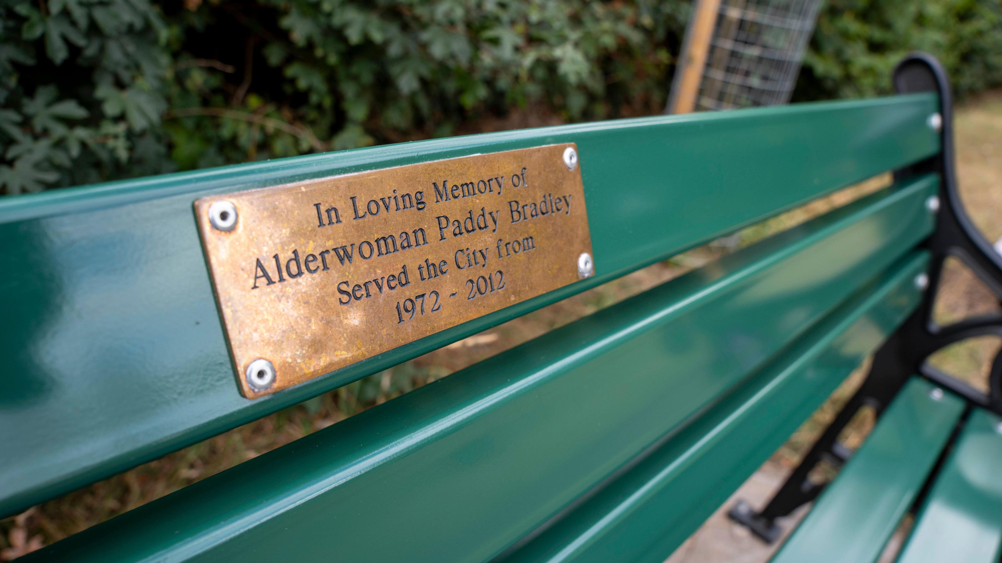 A brass plaque on a green bench reads: "In loving memory of alderwoman Paddy Bradley - served the city from 1972-2012".