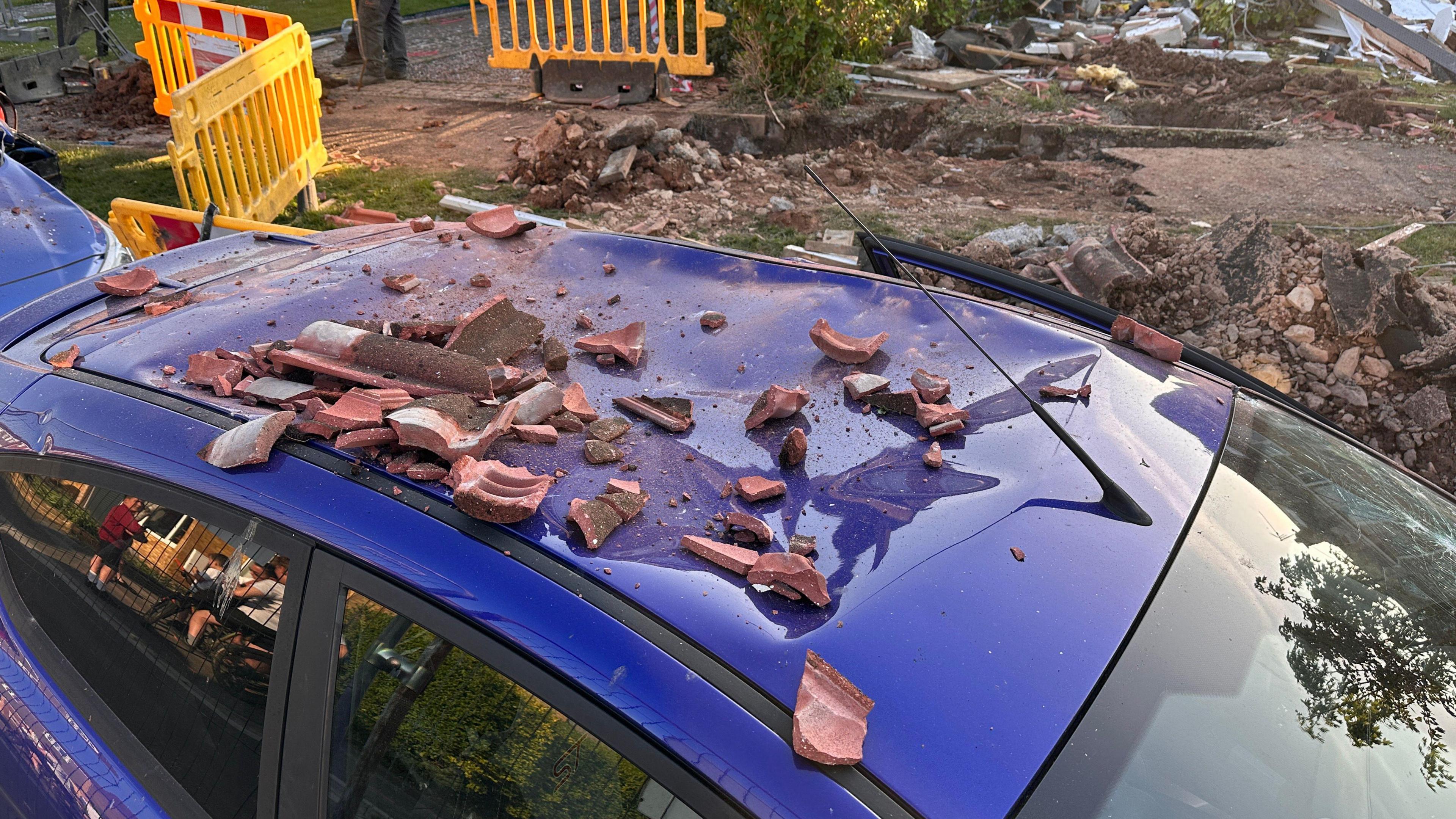 Close up of a car roof which suffered damage in a suspected gas explosion at a house. The car is bright blue and there is debris on the roof. The roof is dented.