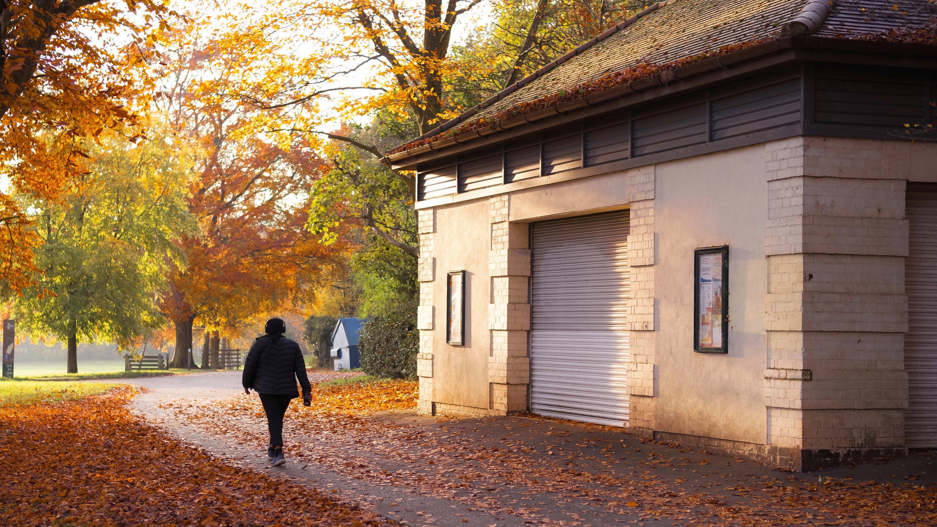 A person wearing headphones walks along a leaf-strewn pathway past a park building towards a row of trees showing autumnal colours