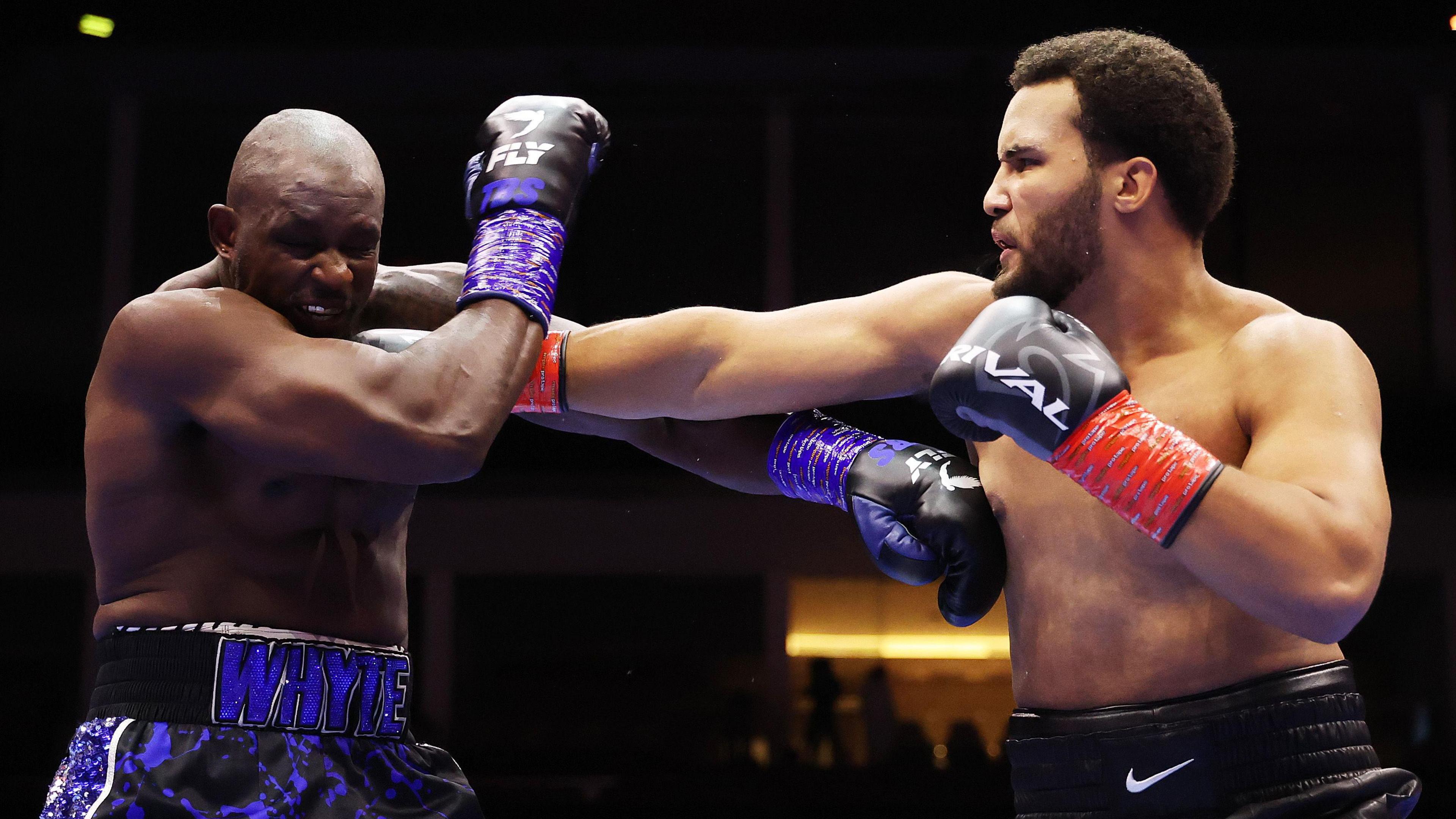 A side-on image shows boxers Moses Itauma and Dillian Whyte from waist up during a fight in August 2025. Itauma is on the right of the image landing a right-handed jab through the middle of Whyte's defences onto his chin, leading his opponent to turn and duck his head towards the camera and screw up his face. The bearded Itauma is wearing black shorts and has black gloves with a red wrist strap, whereas Whyte wears black and purple gloves and shorts in a similar design which also have his surname on the waistband