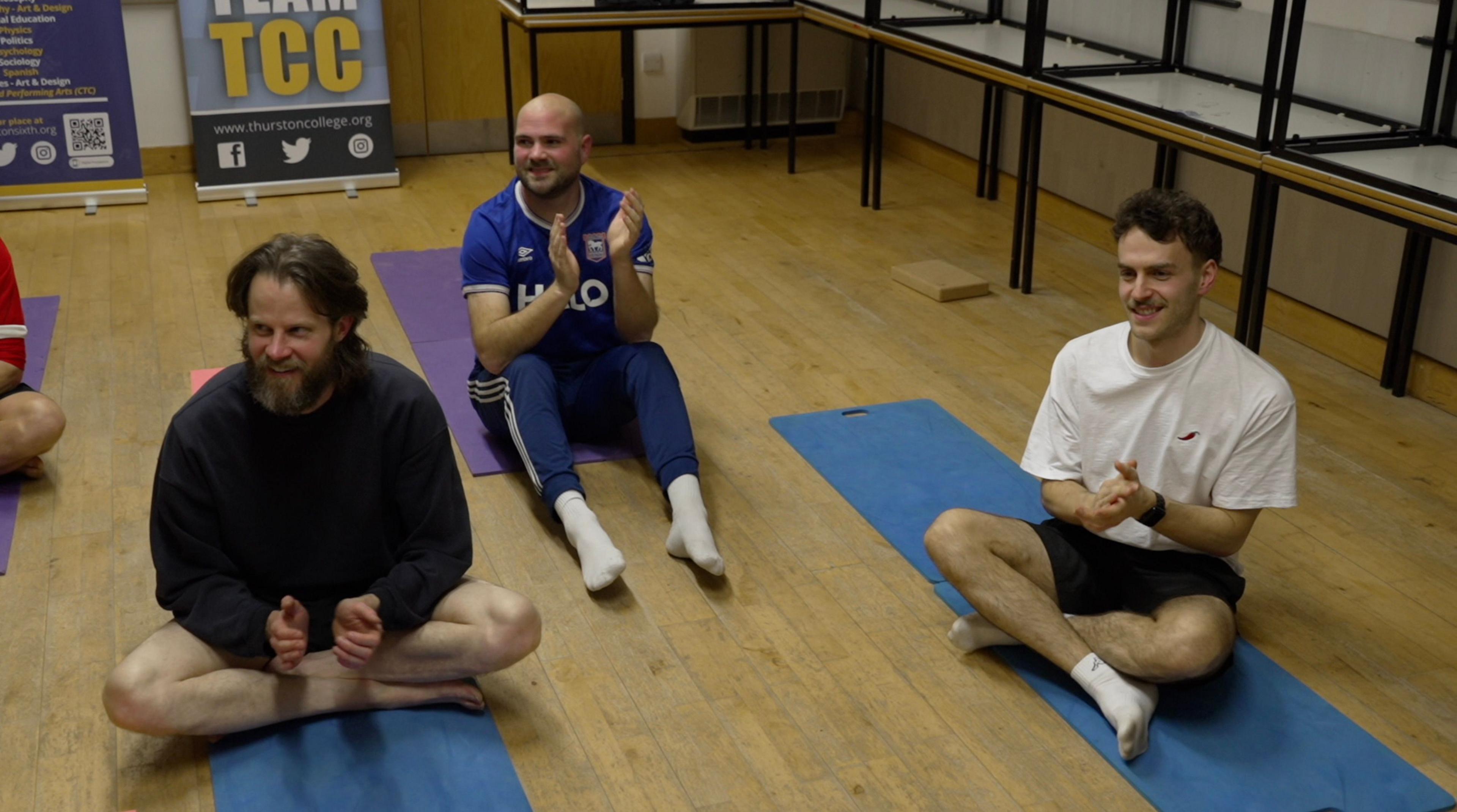 Jack Plumb is sat crossed leg on a yoga mat. He has long hair, and a greying beard. he is wearing shorts and a blue sweater. He is clapping as the session draws to a close. Will Tovey is tot he right, also sat on a blue yoga mat, and clapping.  He is wearing a white T-shirt, has brown hair and moustache. There is also a man between them wearing an Ipswich Town jersey, also applauding.