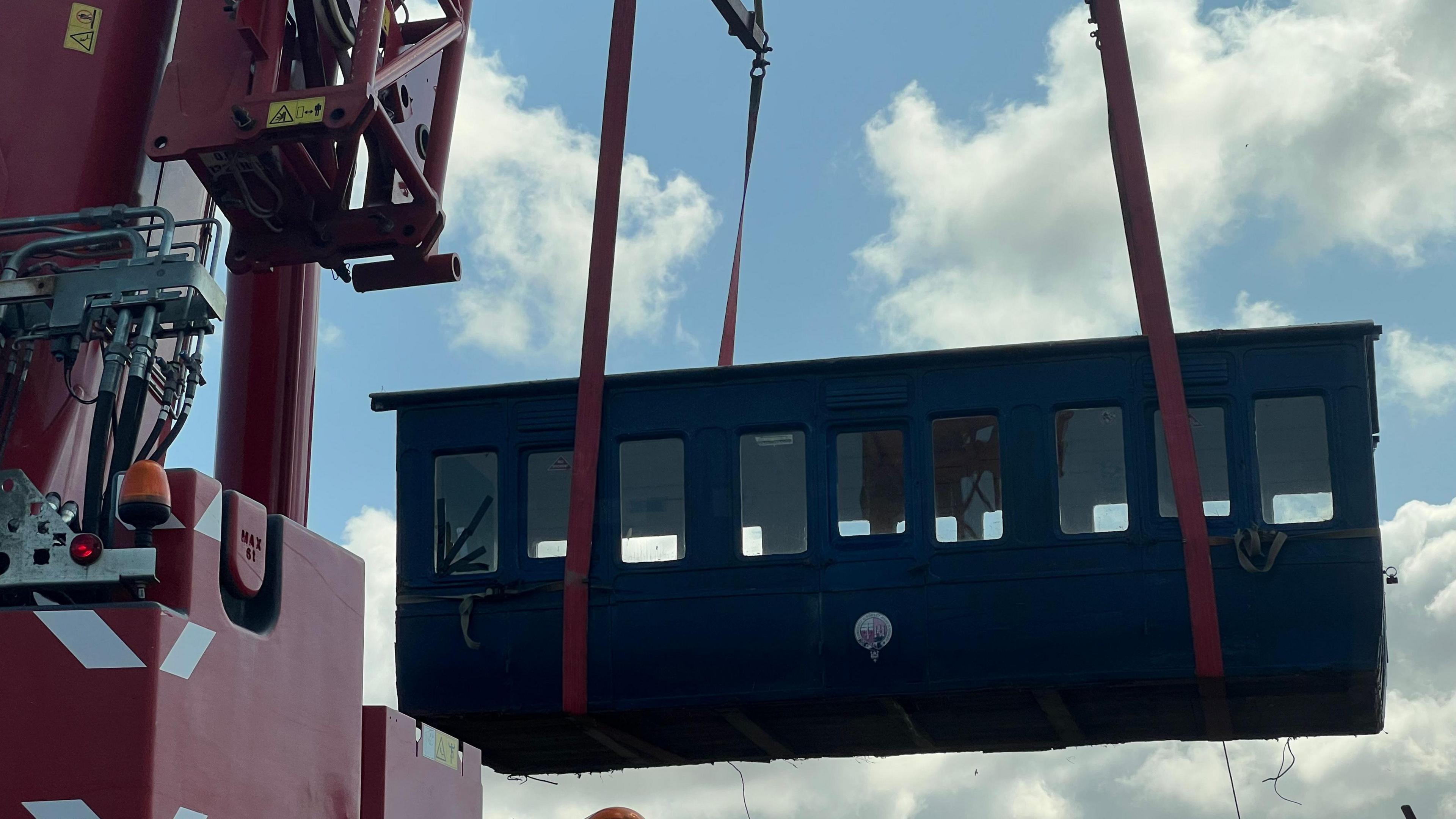 A blue train carriage is being lifted up into a sunny sky by a red crane.