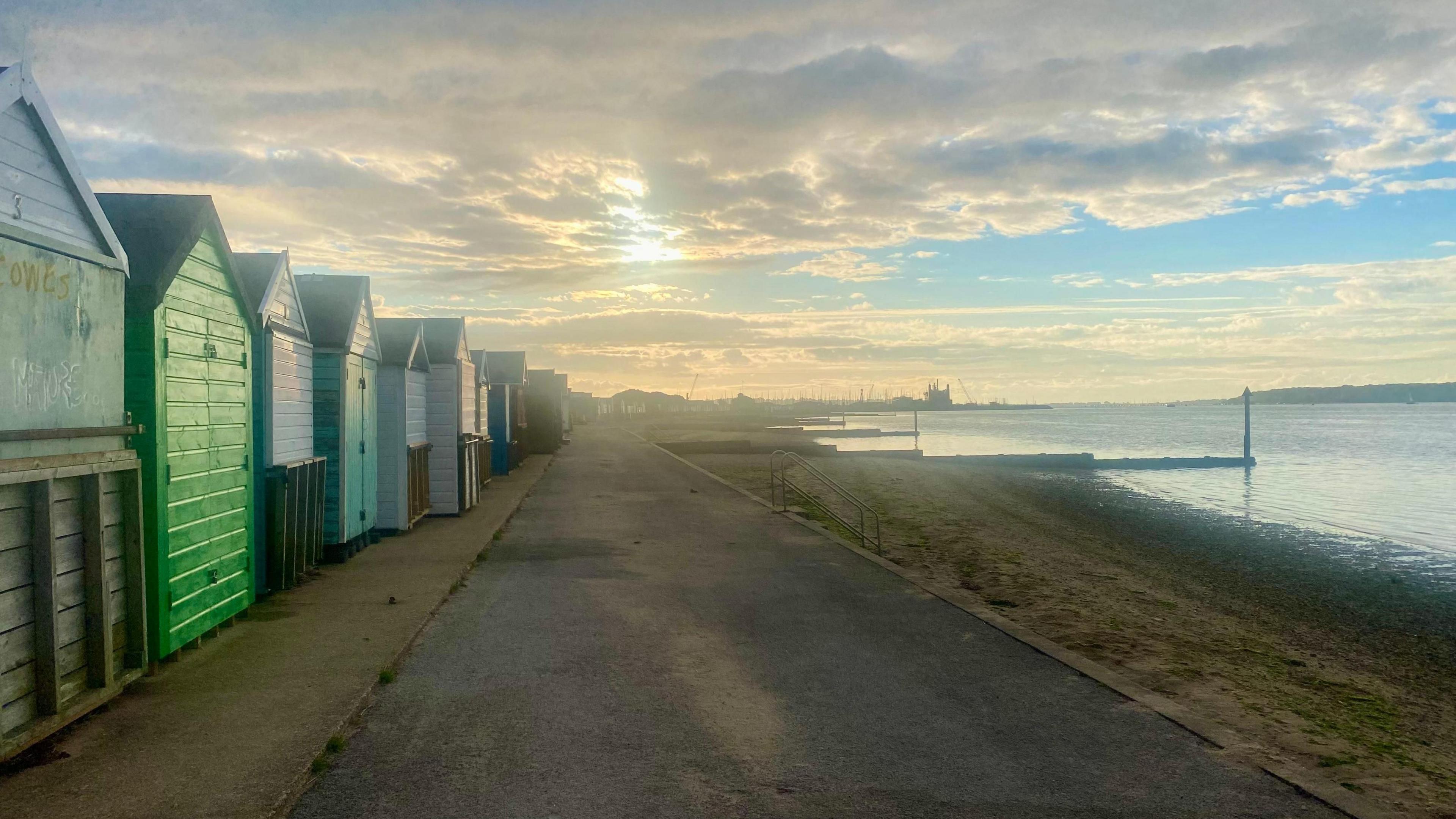 Green, blue and white wooden beach huts on Hamworthy seafront next to a promenade and small beach are lit up by a lowering sun beaming through clouds. 