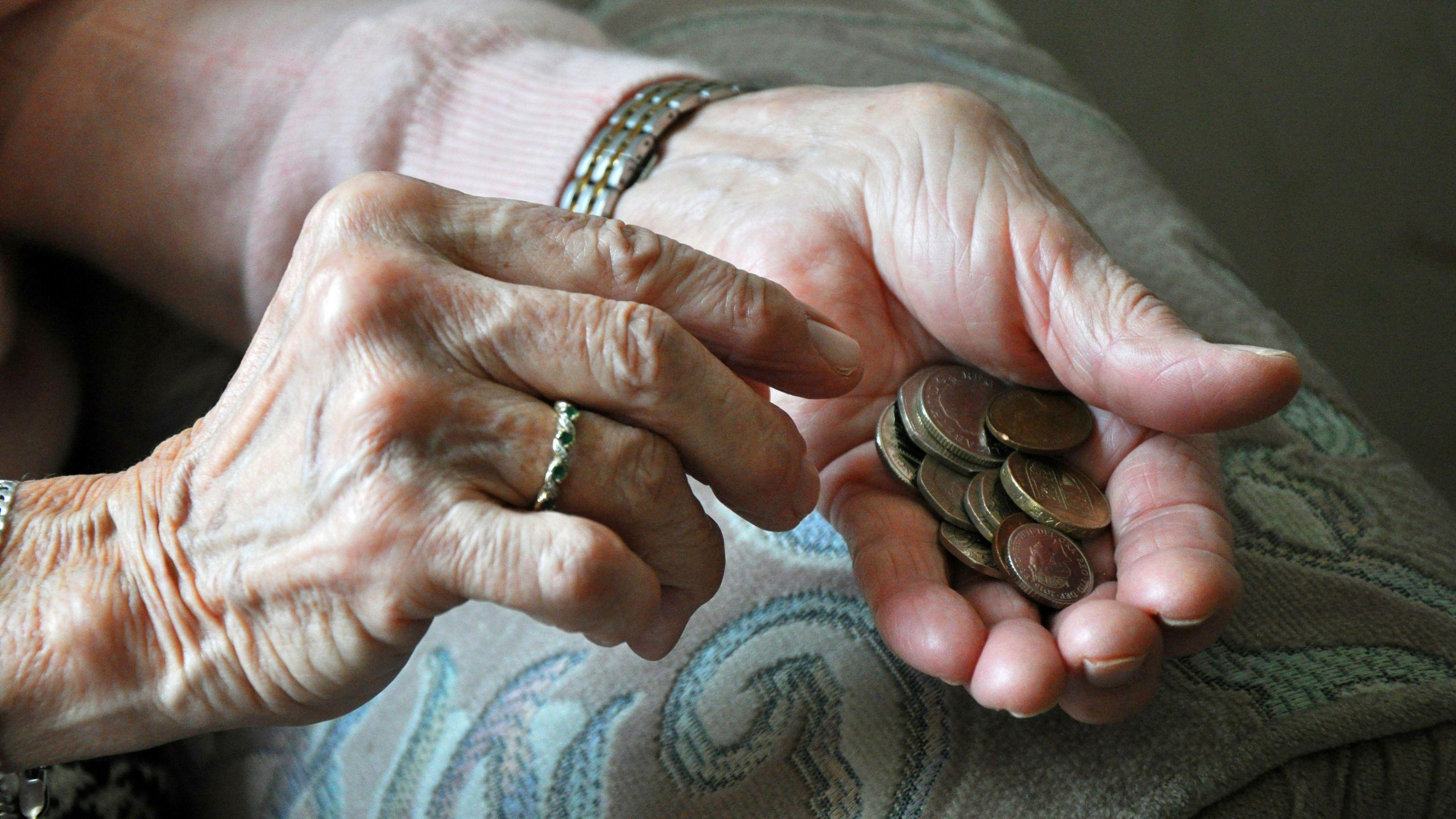 An elderly woman counting loose change. She's wearing a ring and a watch. She's wearing a pink cardigan and is sitting on a sofa.