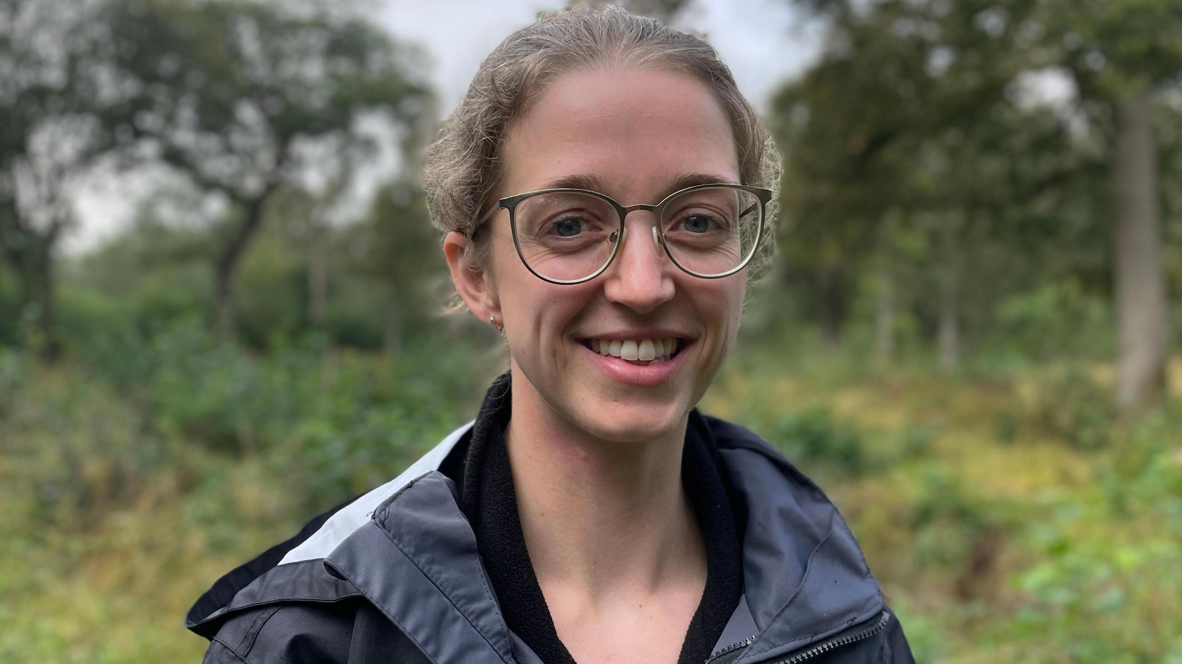 Katharine Flach with pulled back light brown hair, large round brown framed glasses, wearing a blue water proof jacket, standing in Hayley Wood with trees and shrubs behind, smiling