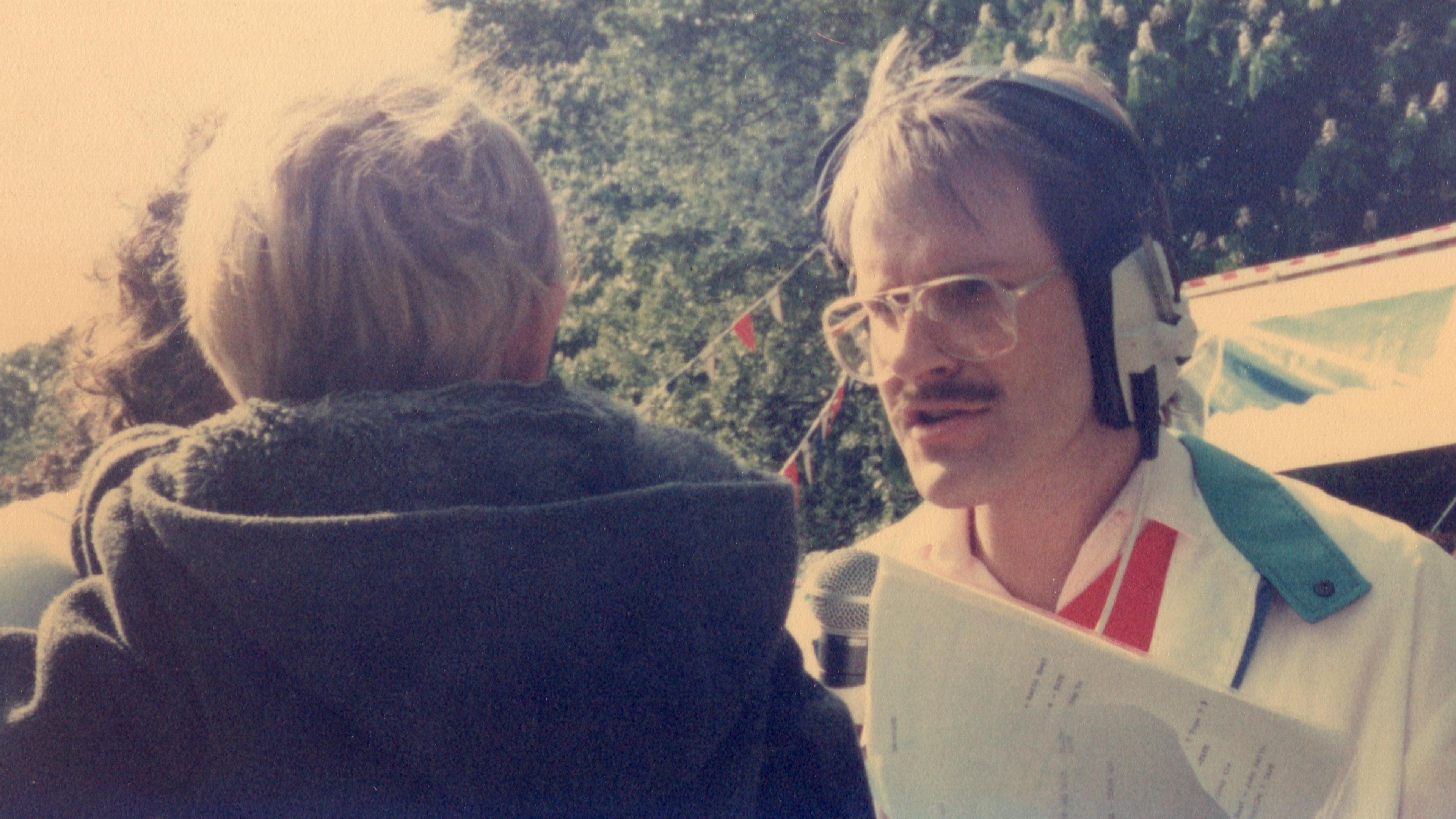 Chris Whitehead wearing a white jacket, headphones and large glasses with thick rims. he is speaking to a person whose face cannot be seen.