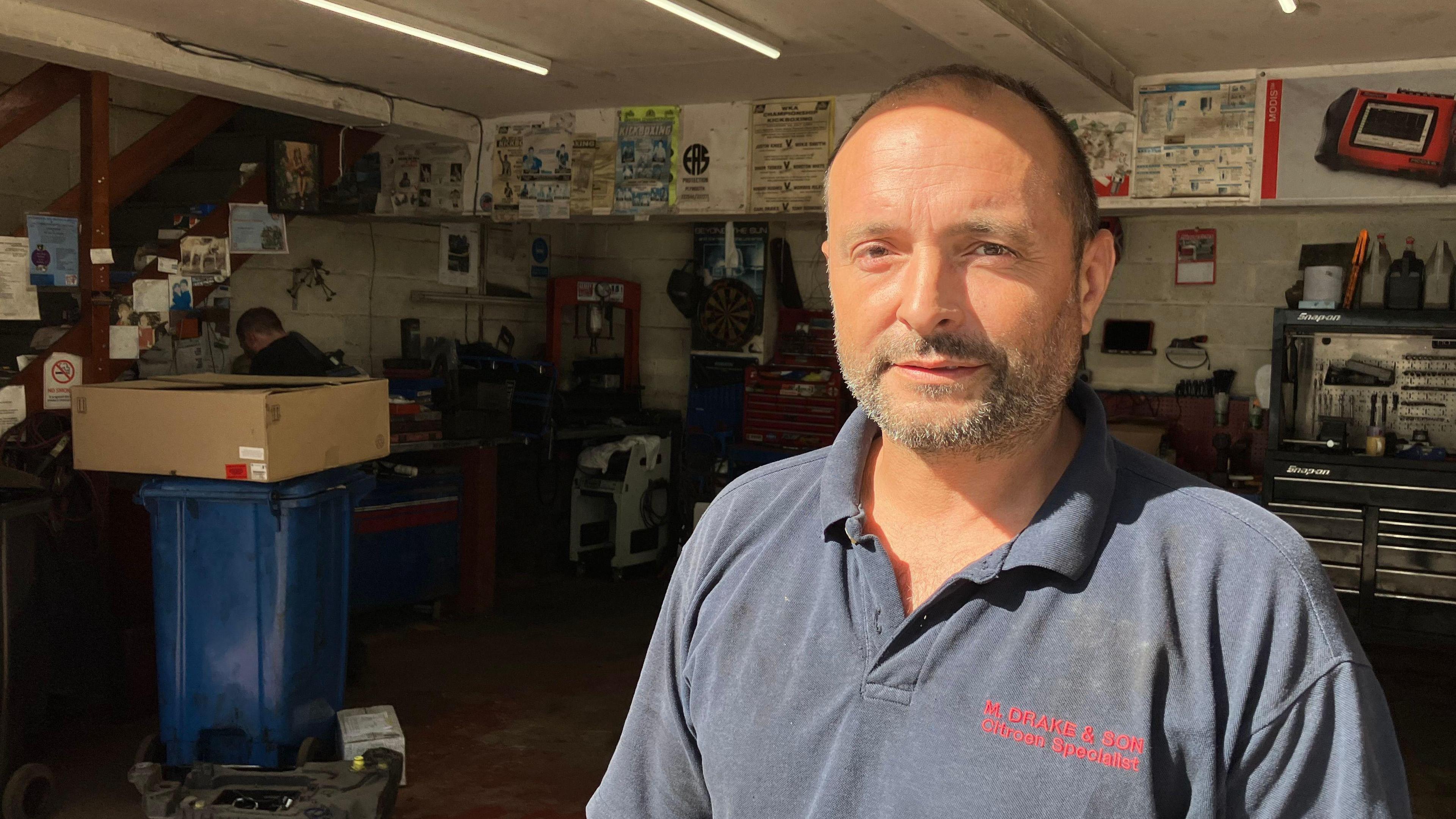 Mechanic Paul Drake, who has short dark hair, a beard and is in a blue T-shirt, stands in the entrance of a garage. There are tools and equipment on the shelves in the background. 