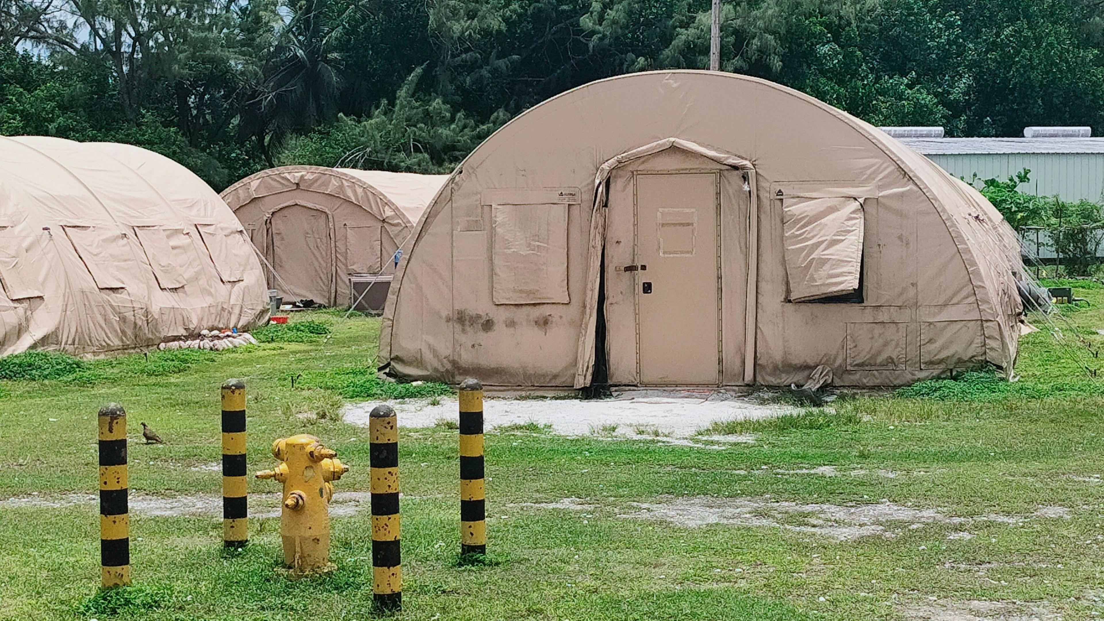 Photo shows three round beige tents on green grass. In the background there is a hut